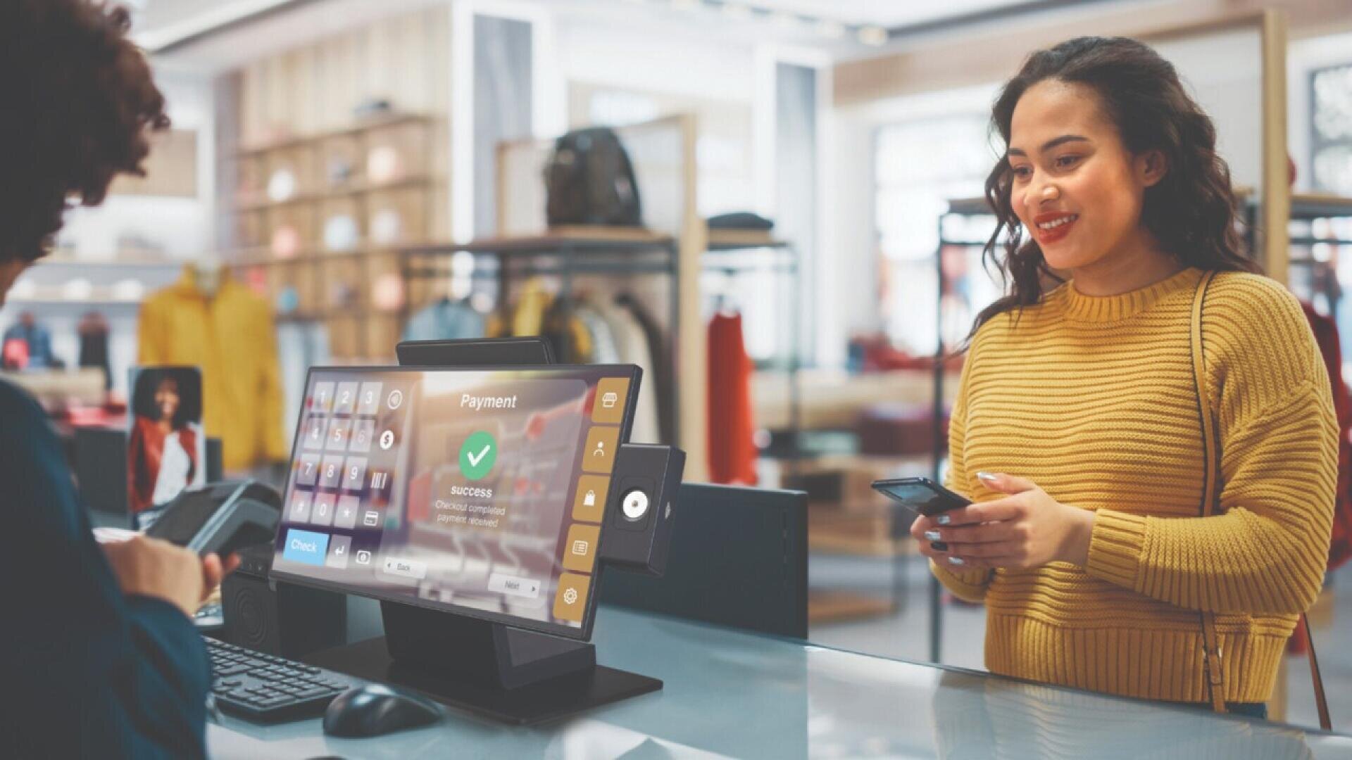 A woman in a yellow sweater smiles while making a payment with her phone at a retail store checkout counter, where a digital screen shows a successful transaction.