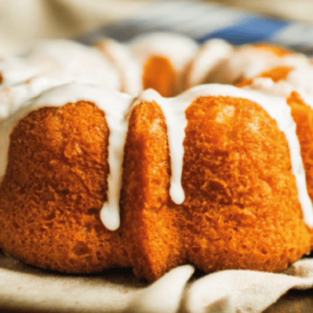 A close-up of a golden-brown bundt cake with white icing drizzled on top, sitting on a beige cloth.