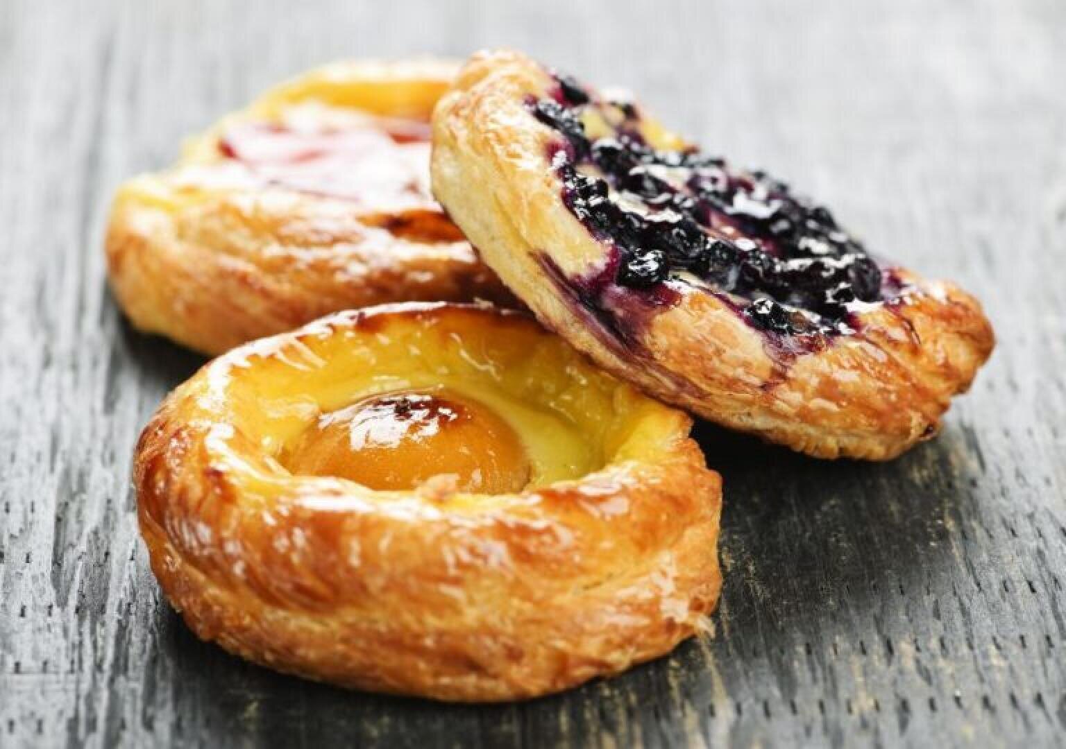 Three round Danish pastries with flaky golden crusts sit on a dark wooden surface. One is topped with blueberry filling, another with apricot, and the third with a red fruit jam.