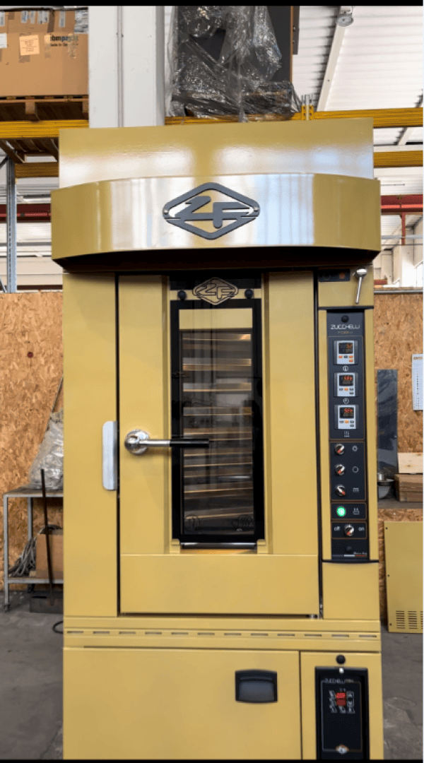 A large yellow industrial bakery oven with a glass door, control panel, and logo on the front stands in a warehouse with shelves and wood paneling in the background.