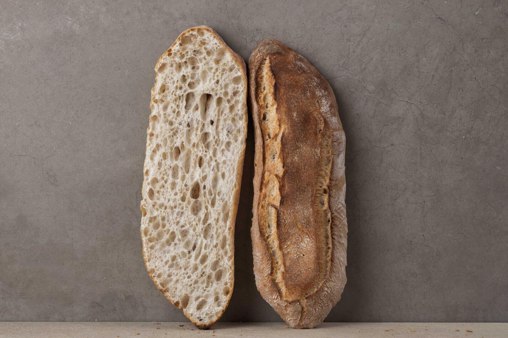A rustic loaf of bread is sliced in half lengthwise and displayed upright, showing a golden-brown crust on one side and an airy, open crumb on the other, against a plain gray background.