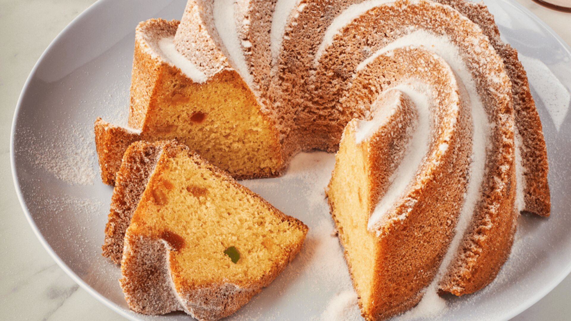 A Bundt cake dusted with powdered sugar is on a white plate, with two slices cut. Visible pieces of candied fruit are inside the cake. In the background, a plate with a cake slice and pink glasses are on a marble surface.