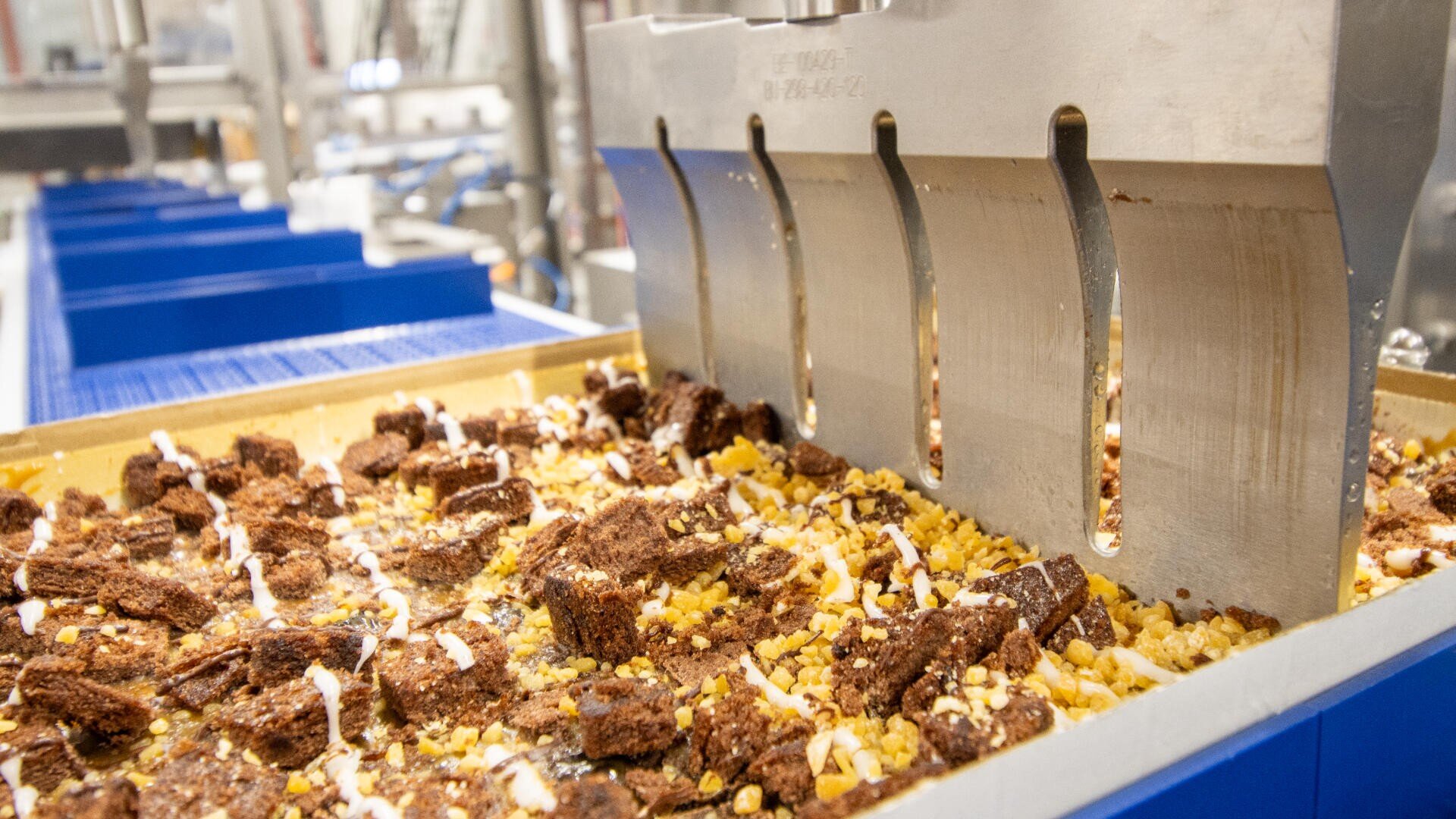 A close-up of a food processing machine slicing a tray of dessert bars with chocolate chunks, nuts, and drizzle, inside a factory setting.