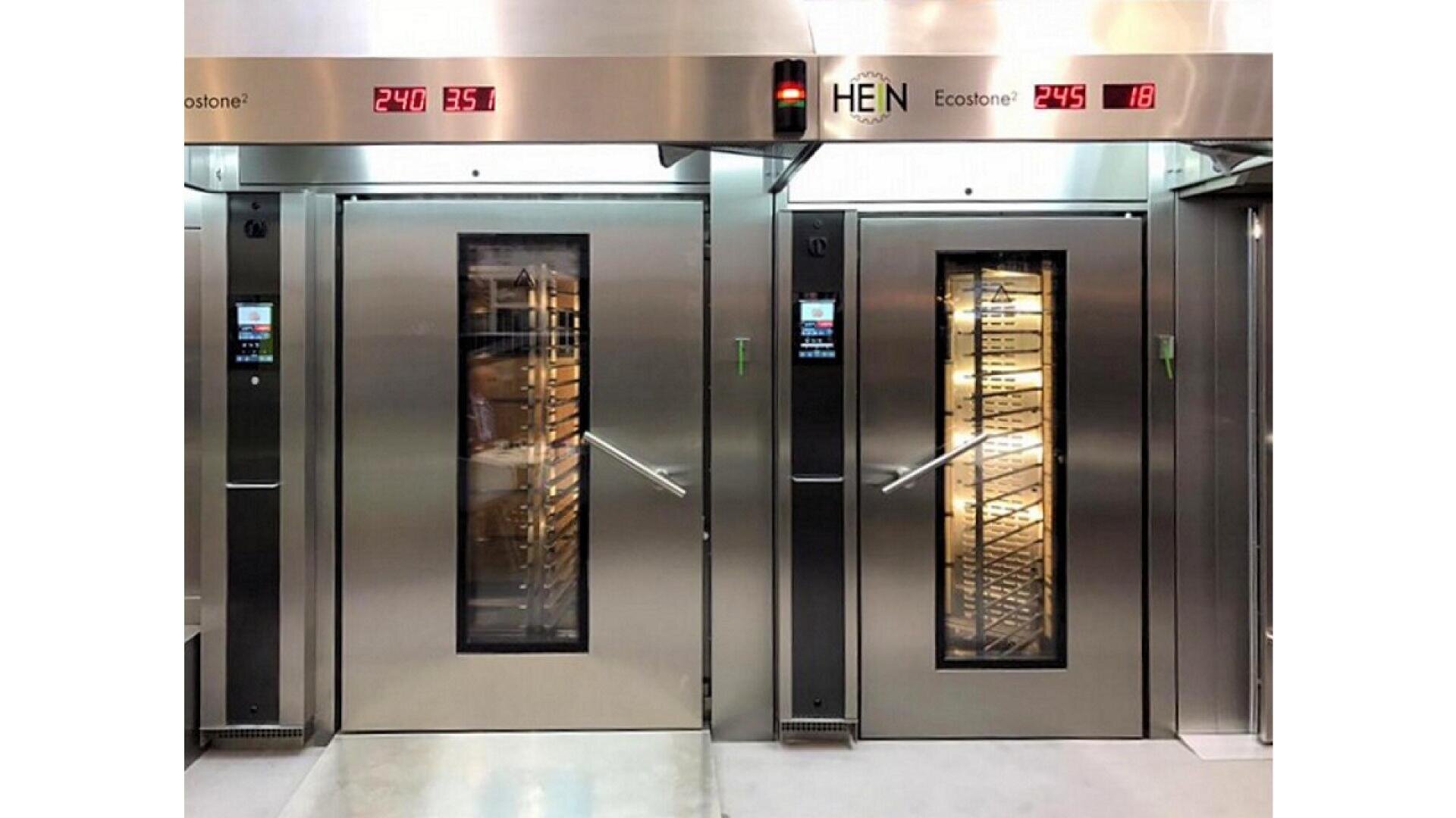 Two industrial stainless steel bakery ovens with digital displays, glass doors, and racks of trays visible inside. The ovens show temperature readings above the doors. The setting appears clean and modern.