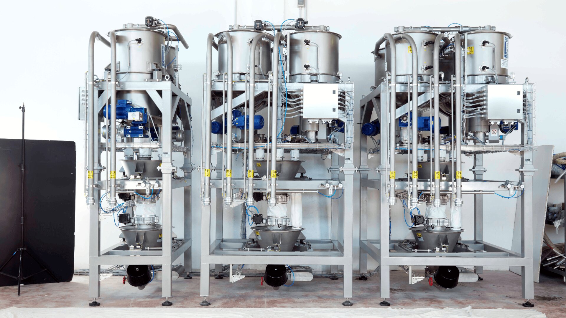 Three large, stainless steel industrial machines with tanks, pipes, and control panels stand side by side in a clean, bright facility. The equipment features blue and yellow components and is elevated on metal frames.