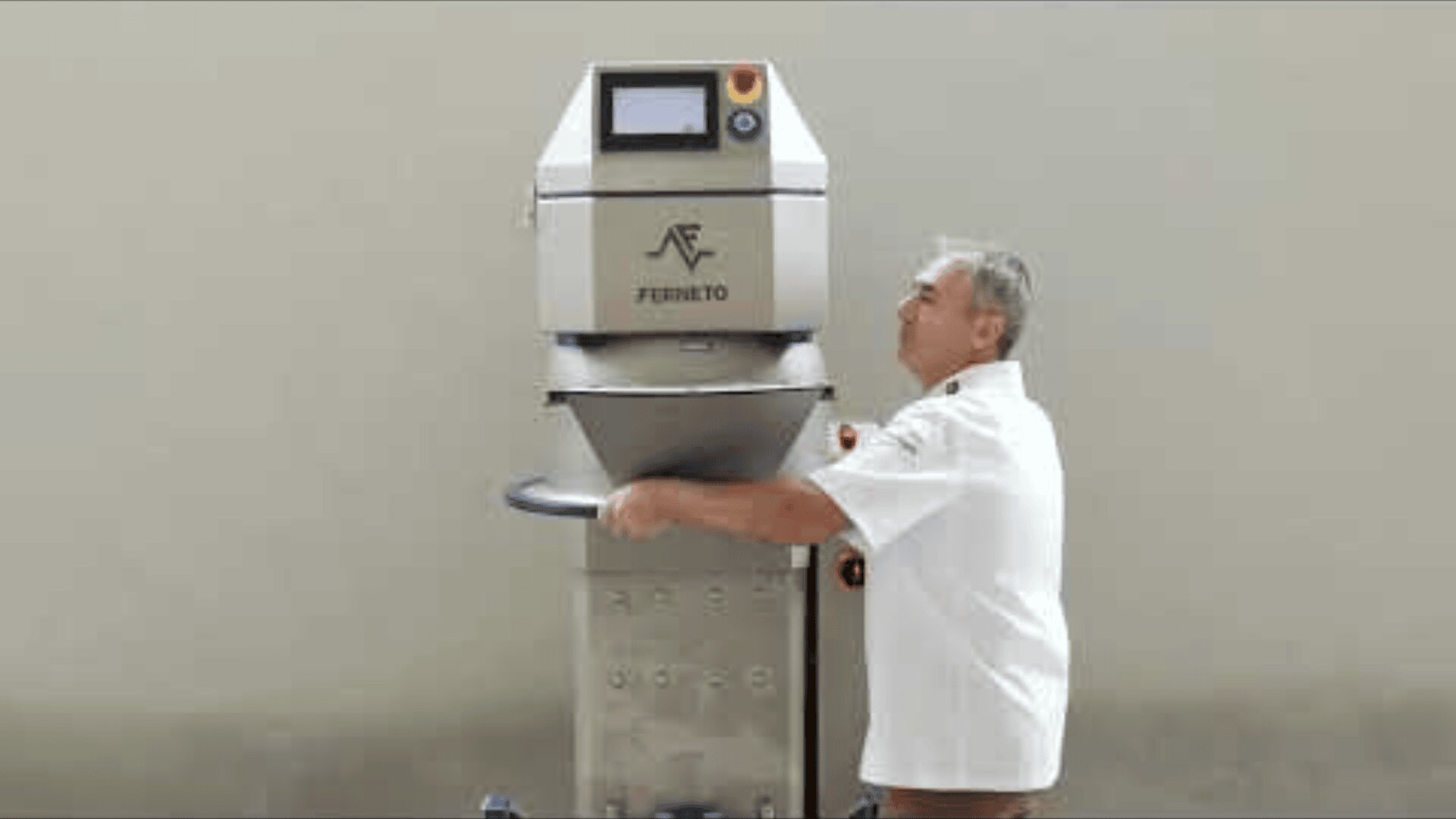 A man in a white shirt operates a large, industrial dough kneading machine labeled Ferneto, which stands against a plain, light-colored wall.