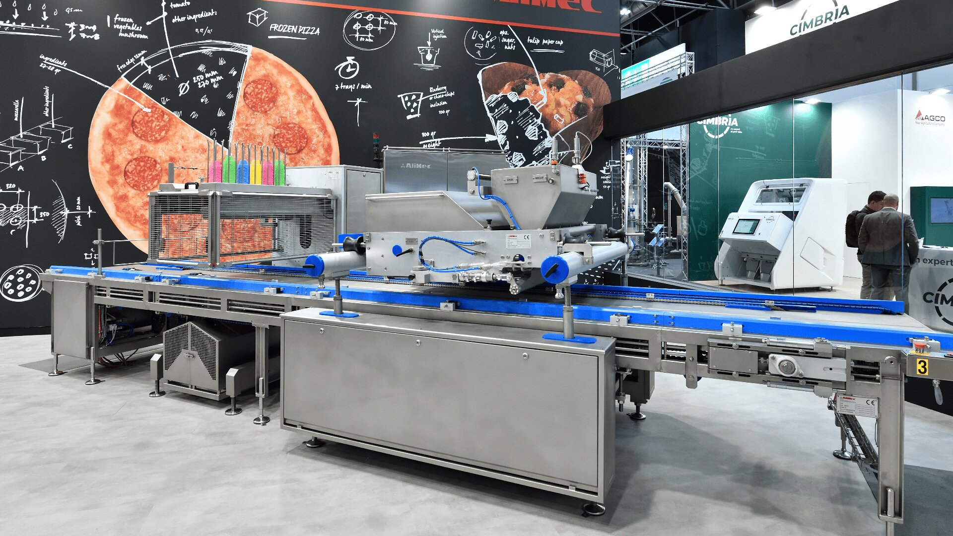 A modern industrial pizza production machine with conveyor belts on display at a trade show, set against a backdrop featuring pizza illustrations and scientific diagrams.