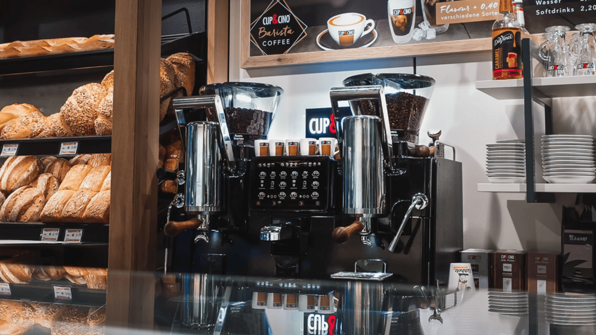 Eine moderne Kaffeemaschine steht auf einer Theke in einer Bäckerei, umgeben von Broten in Regalen und Stapeln von Kaffeetassen. In den Trichtern der Maschine sind Kaffeebohnen zu sehen, und in der Nähe sind verschiedene Getränke und Zubehör ausgestellt.