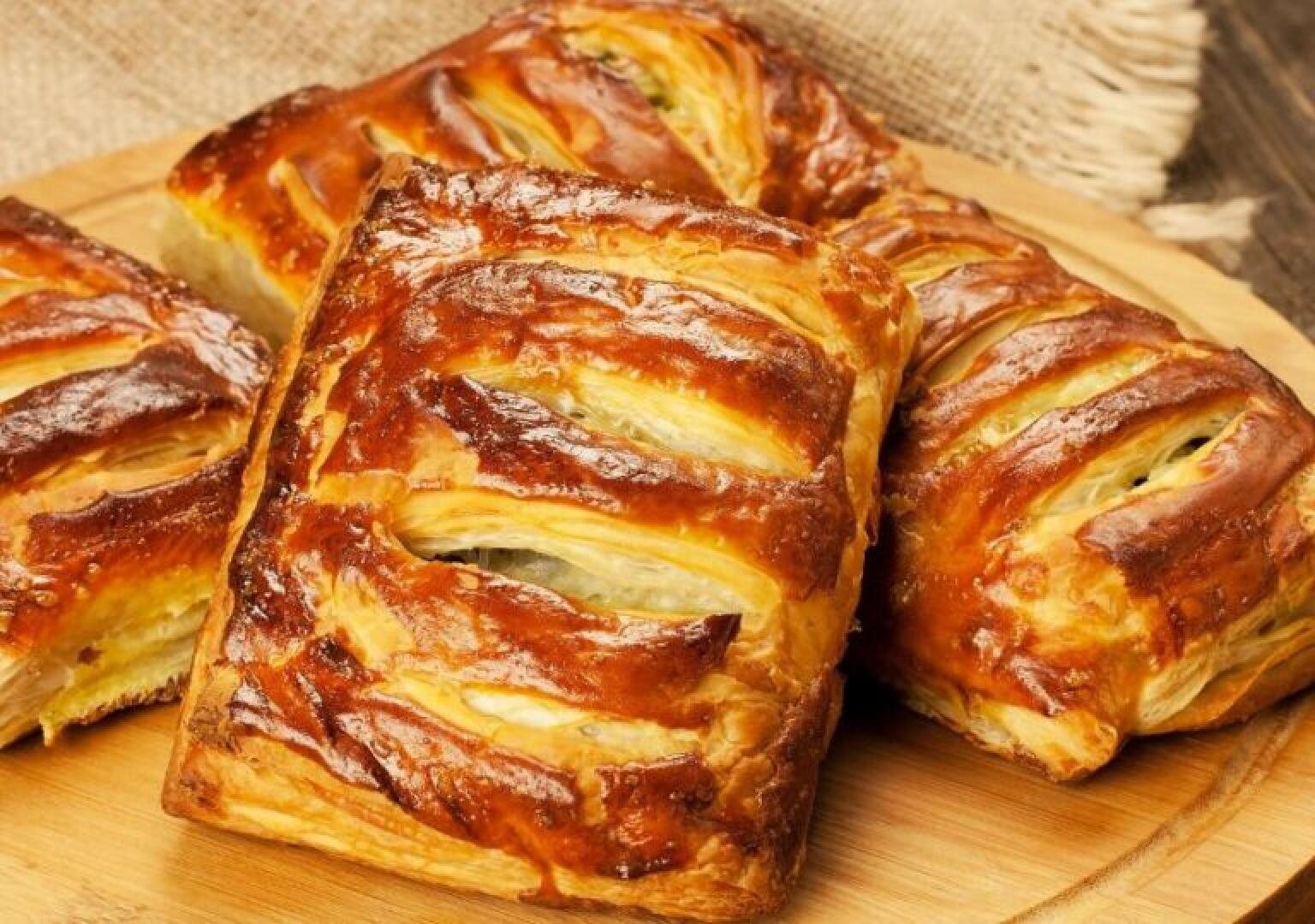 Golden-brown puff pastries with flaky, glazed tops and visible layers are arranged on a wooden board, suggesting a freshly baked, appetizing treat.