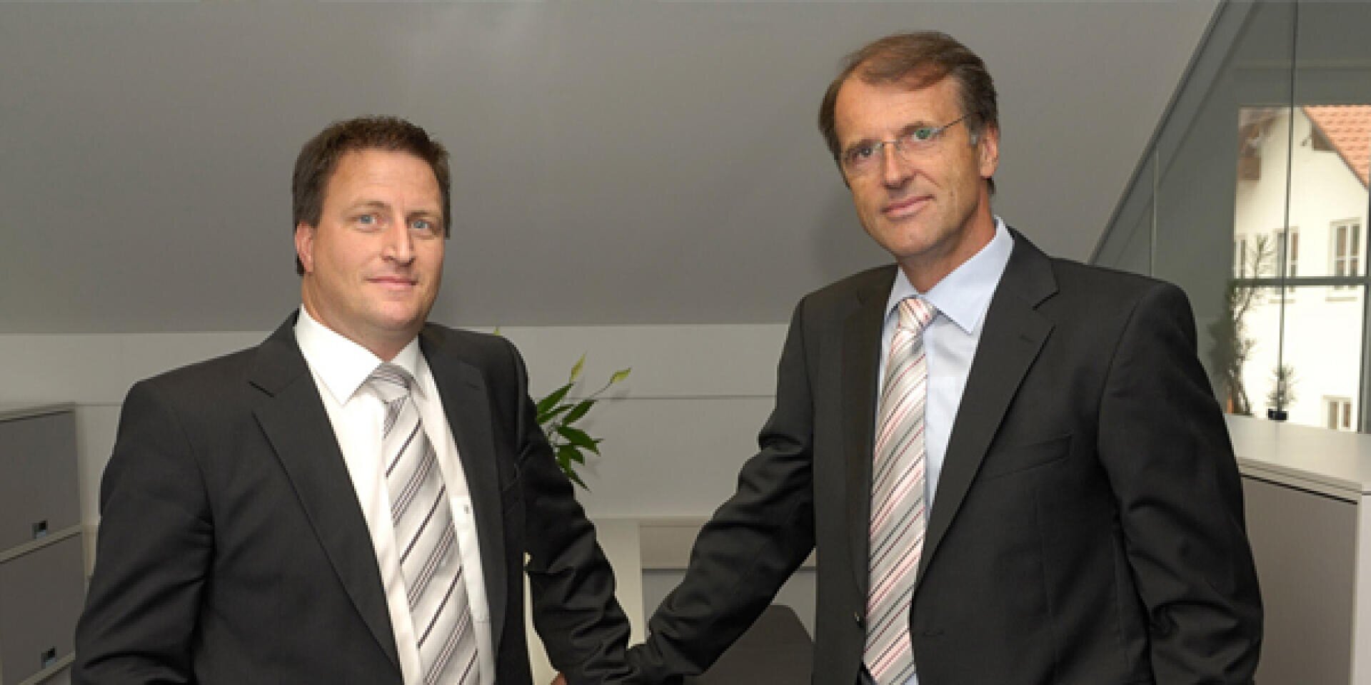 Zwei Männer in Geschäftsanzügen stehen nebeneinander in einem Büro. Beide tragen Krawatten und haben kurze Haare. Der eine Mann steht mit der Hand auf einem Schreibtisch, im Hintergrund stehen Schränke und eine Pflanze.