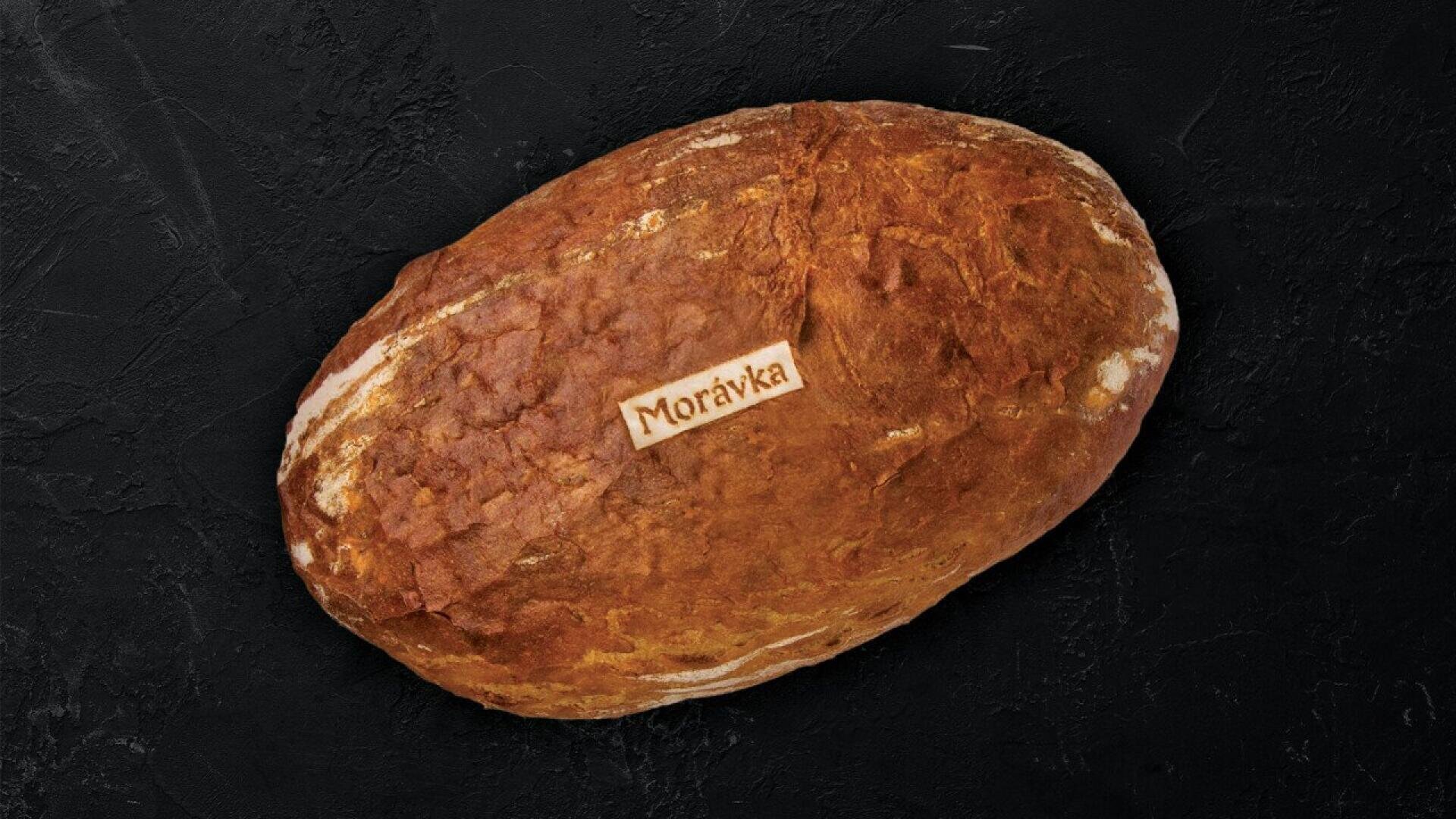 A round loaf of brown bread with a light rectangular label reading Morávka placed on top, set against a dark textured background.