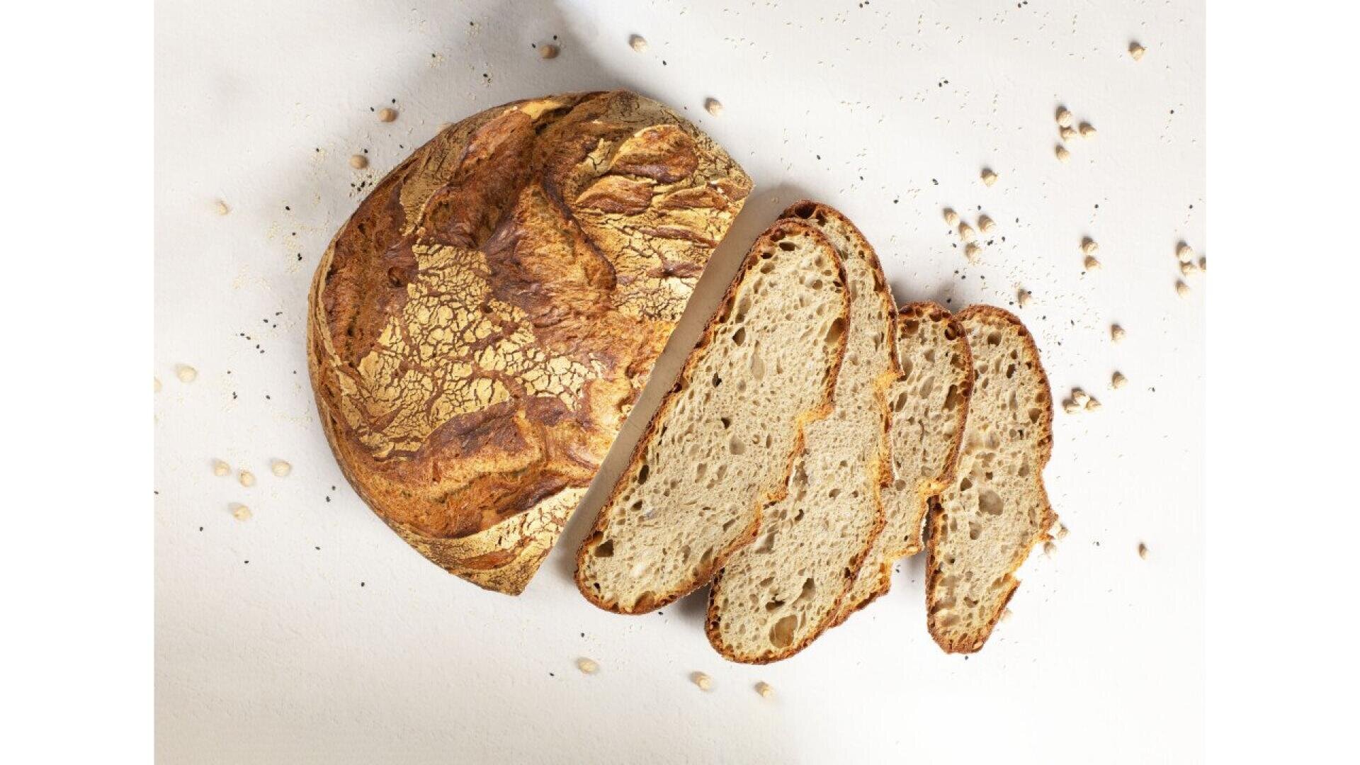 A round loaf of rustic bread, partially sliced, with three slices laid out on a white surface. There are scattered crumbs and grains around the bread.