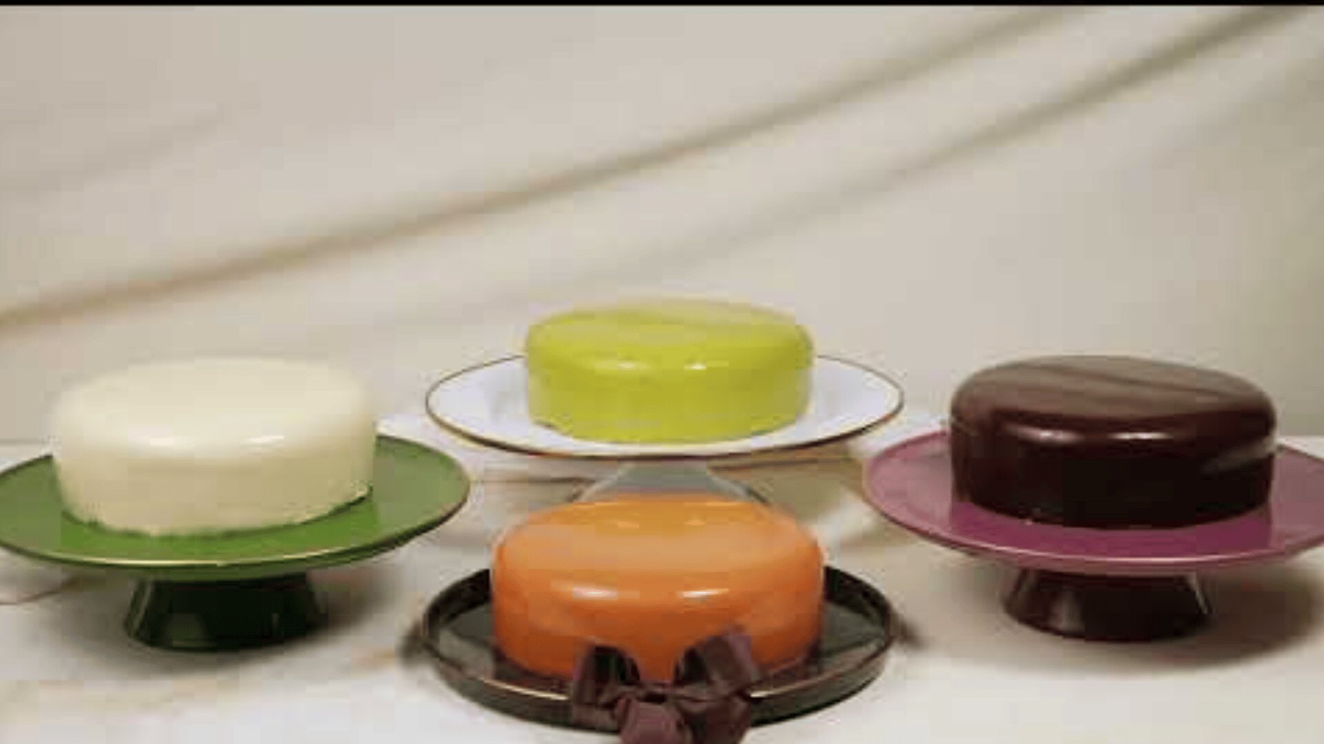 Vier runde Torten mit glatter, glänzender Glasur in Weiß, Grün, Orange und Dunkelbraun werden auf verschiedenen Tortenständern vor einem hellen, drapierten Stoffhintergrund präsentiert.