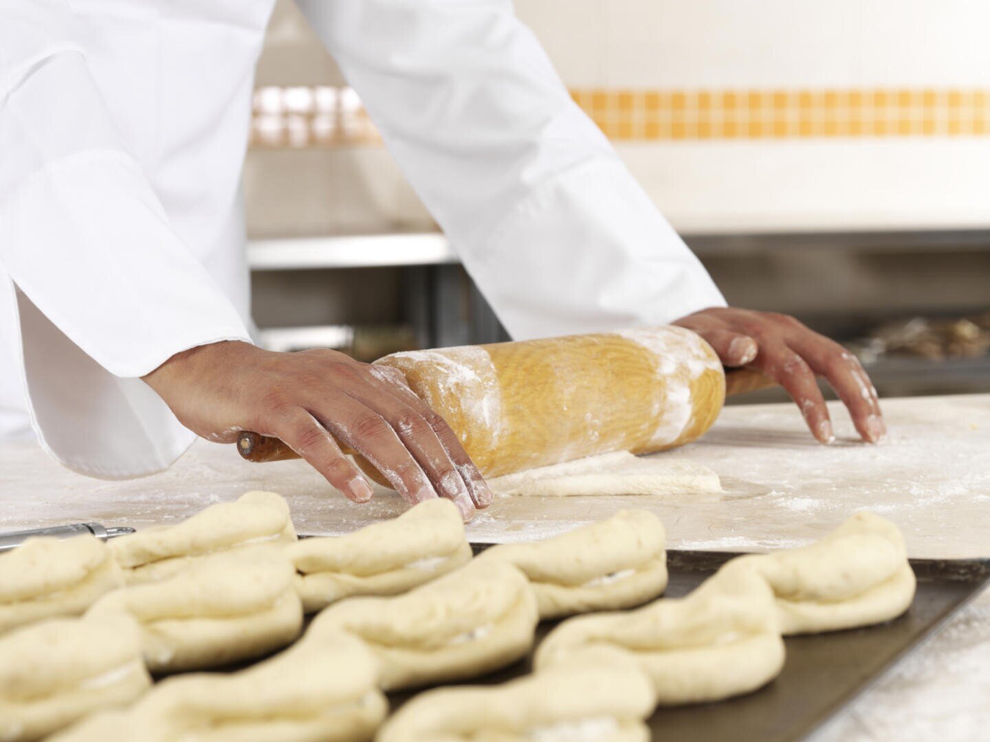 A person in a white chefs coat uses a rolling pin to flatten dough on a floured surface, with rows of shaped dough pieces on a tray in the foreground.