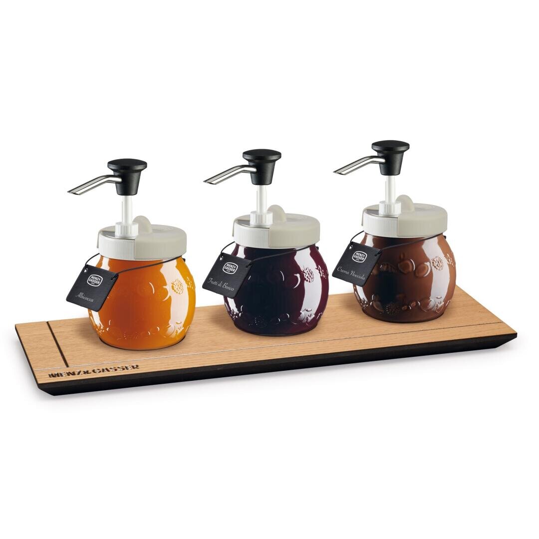 Drei Gläser mit weißen Deckeln und schwarzen Pumpspendern, gefüllt mit verschiedenfarbigen Saucen oder Sirupen, stehen auf einem Holztablett. Jedes Glas hat ein schwarzes Etikett auf dem Deckel.