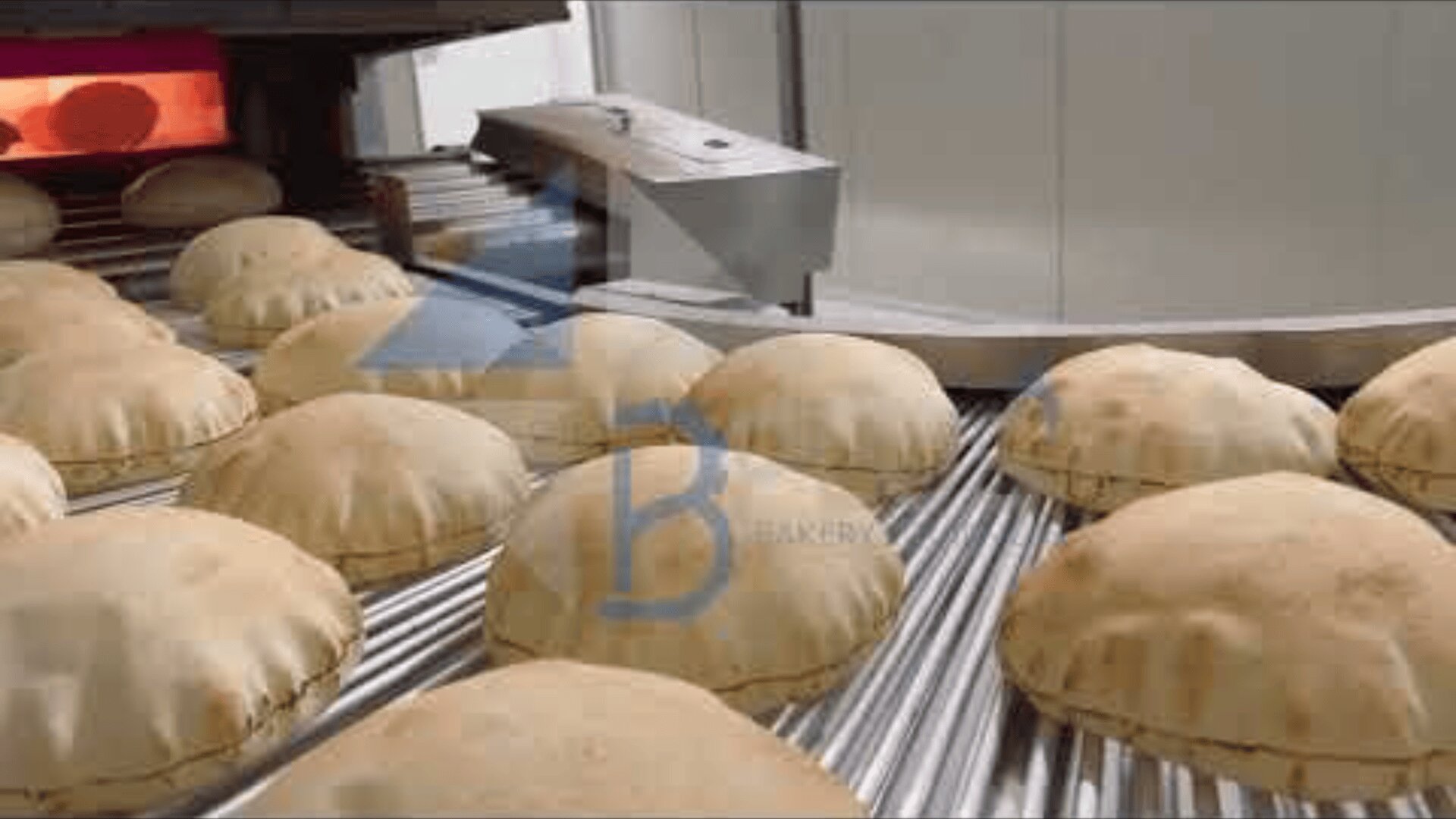 Rows of freshly baked pita bread are moving on a metal conveyor belt inside a commercial bakery, with an industrial oven visible in the background.