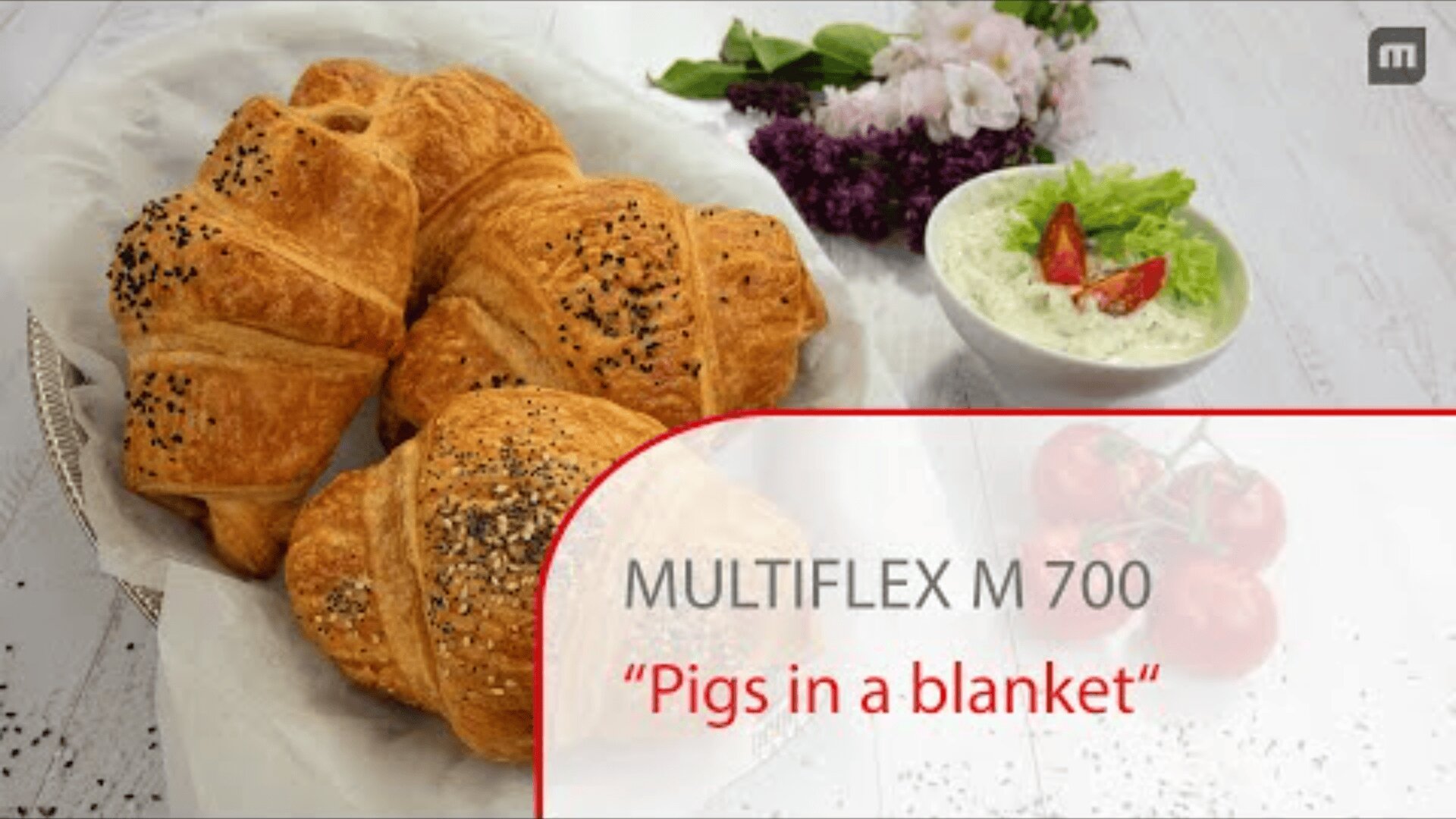 Ein Teller mit goldgelben, flockigen Hörnchen mit Sesam und Mohn, daneben eine Schüssel Salat mit Salat und Tomate, im Hintergrund lila Blumen. Der Text lautet: MULTIFLEX M 700 Würstchen im Schlafrock.