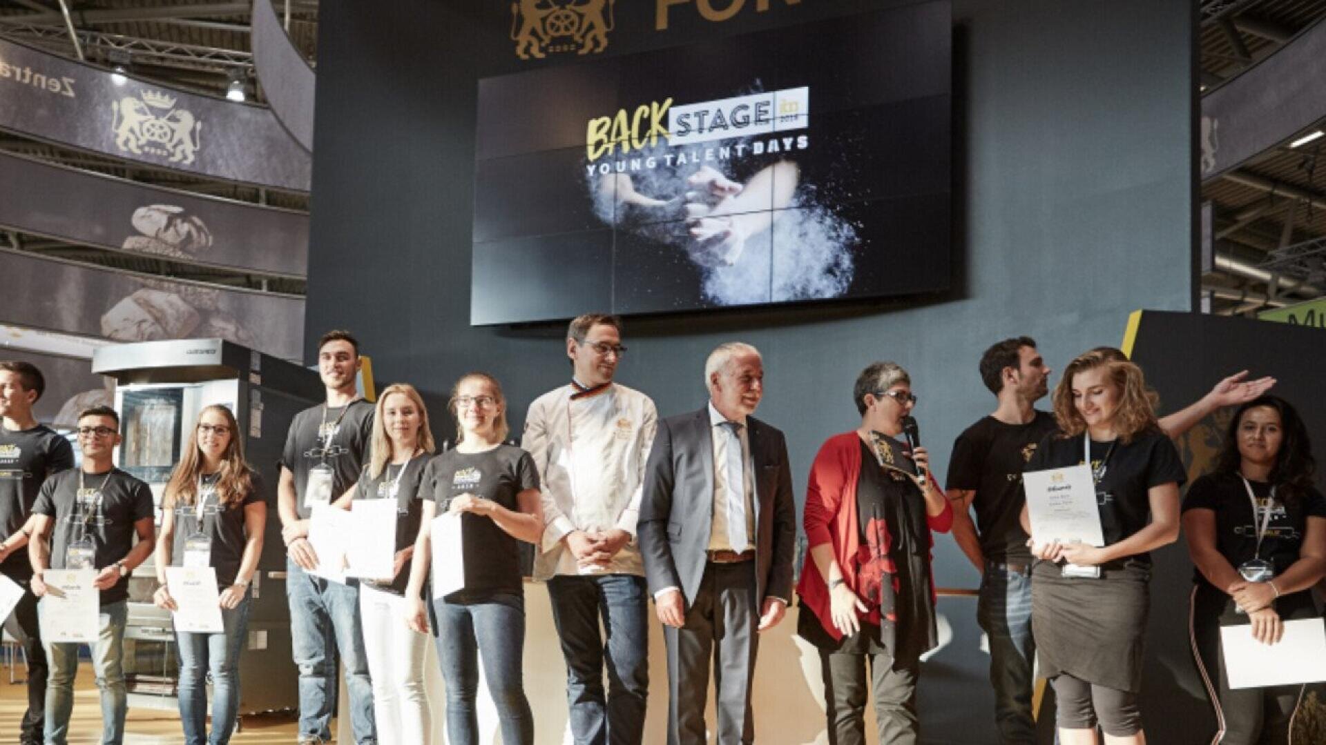 A group of people stand on a stage holding certificates, with a few officials in the center. A large screen behind them displays Backstage Young Talent Days. The setting appears to be an indoor event or competition.