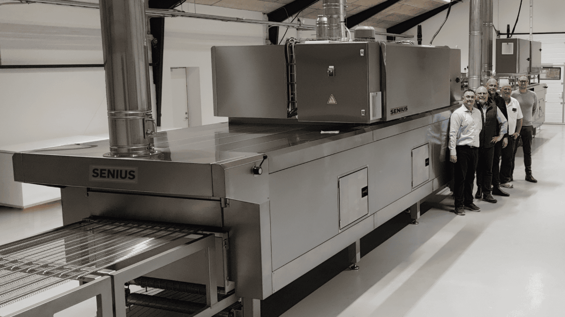 Five men in business attire stand beside a large industrial oven labeled SENIUS in a clean, modern facility. The oven has a metal conveyor belt and prominent ventilation pipes.
