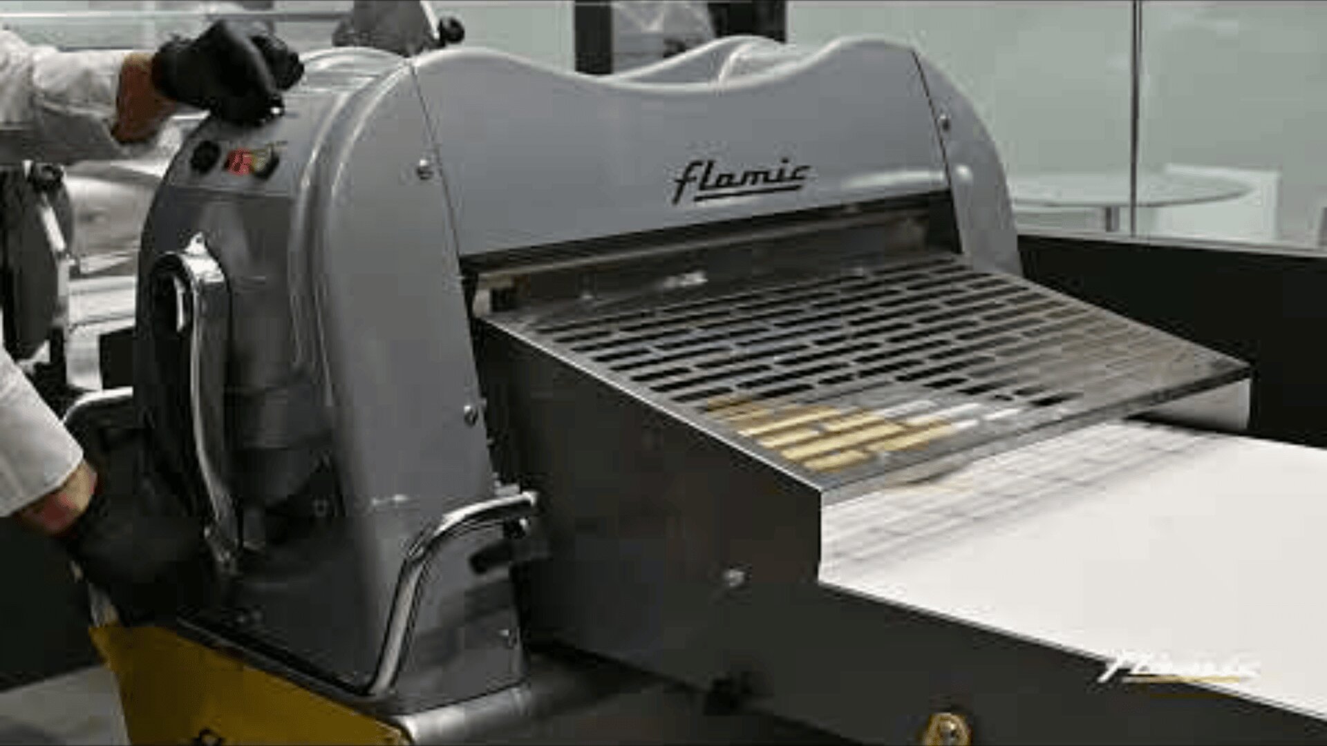 Eine Person bedient eine große industrielle Teigausrollmaschine mit der Bezeichnung Flamic, wobei der Teig durch Walzen auf ein Förderband geleitet wird.