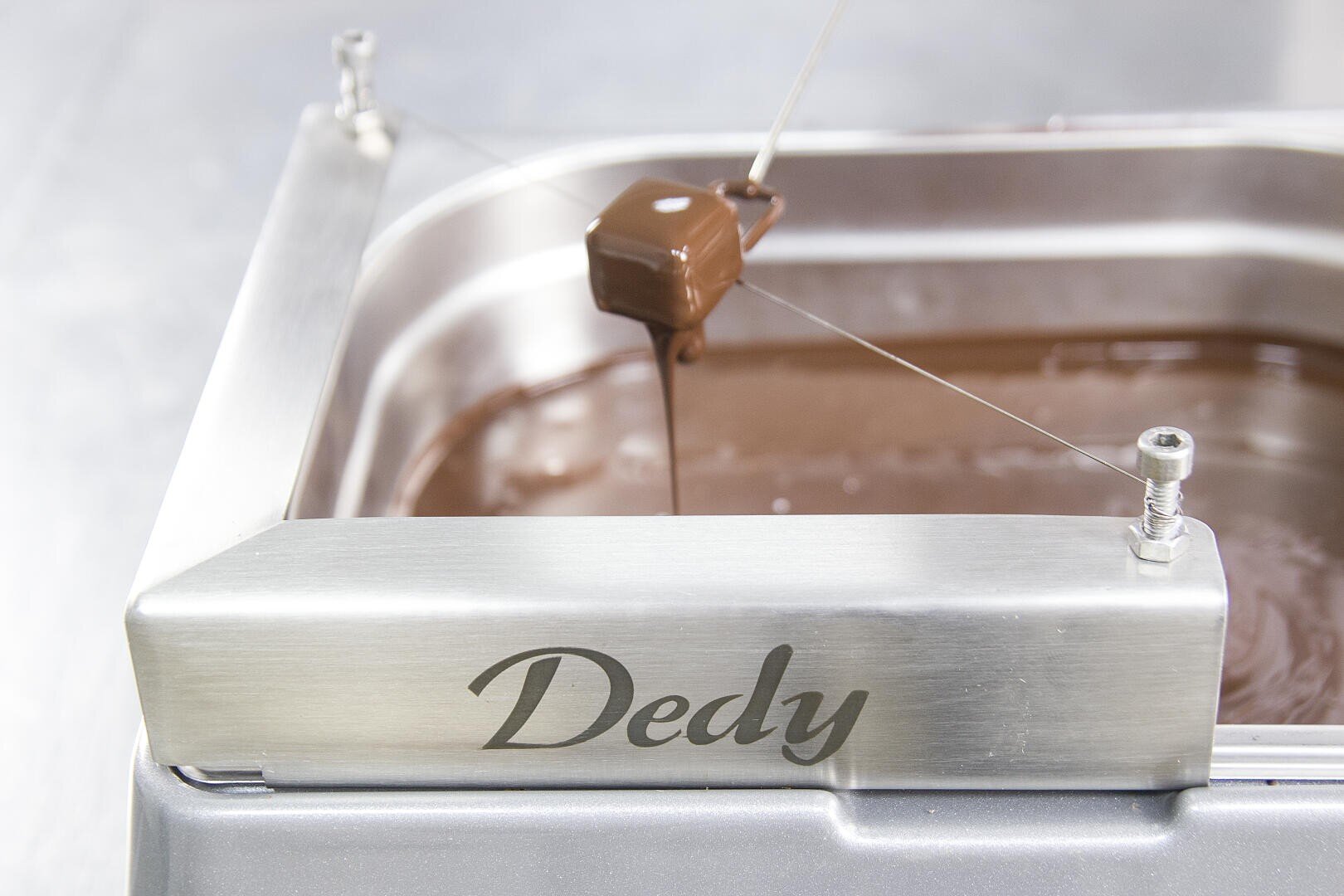Ein Stück schokoladenüberzogenes Konfekt wird von einer Tauchgabel über einem Behälter mit geschmolzener Schokolade gehalten, wobei Dedy auf dem Metallrand der Maschine eingraviert ist.