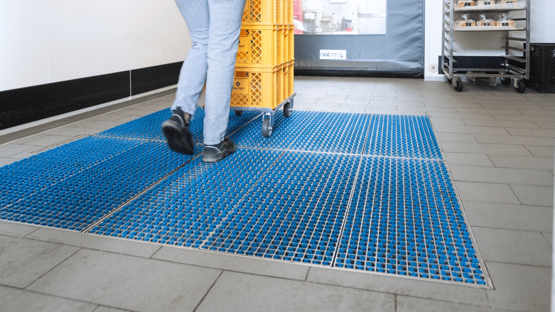 Eine Person schiebt einen Wagen mit gelben Kisten über blaue Bodenmatten mit Metallrosten in einem sauberen Innenraum, möglicherweise in einer Großküche oder einer Lebensmittelfabrik.