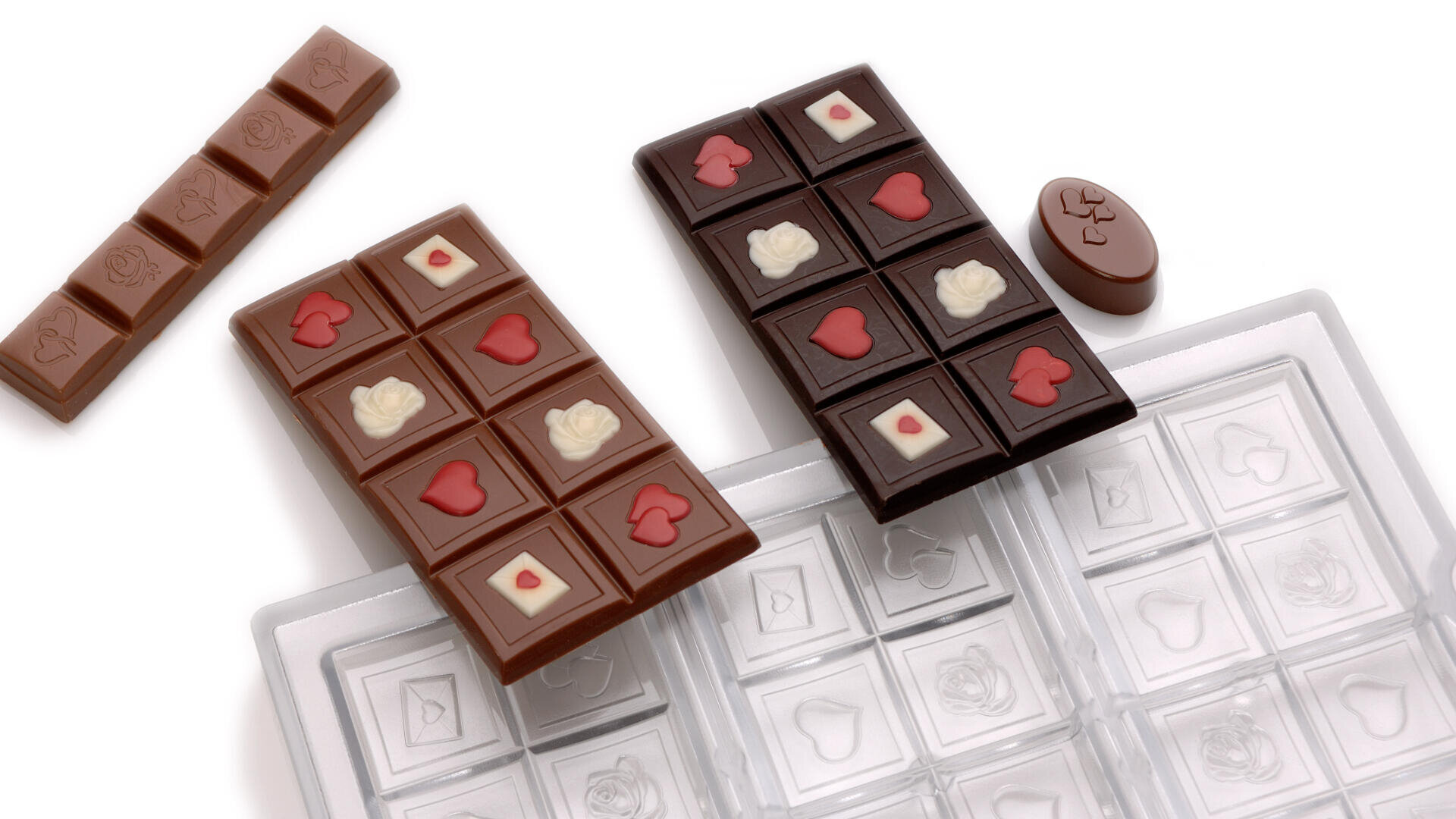 Zwei Schokoladentafeln mit Herz- und Rosendekor, eine aus Vollmilch- und eine aus Zartbitterschokolade, liegen neben einer leeren Schokoladenform. Eine kleine, ovale Schokolade mit Herzformen steht daneben, und ein Schokoladenstück liegt waagerecht oben links.