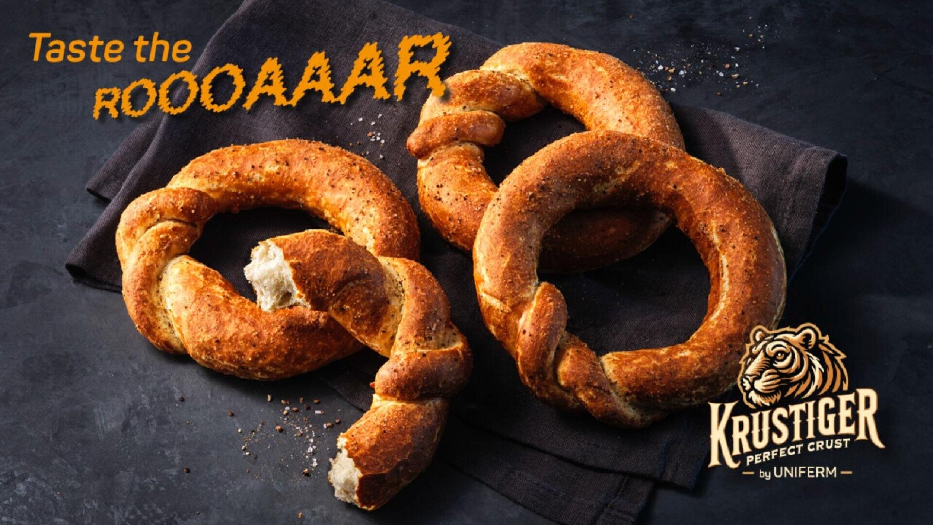 Three golden, twisted bread rings sit on a dark cloth, with one partially eaten. Bold text reads Taste the ROOOAAAR, and a tiger logo with Krustiger Perfect Crust by Uniferm appears in the corner.