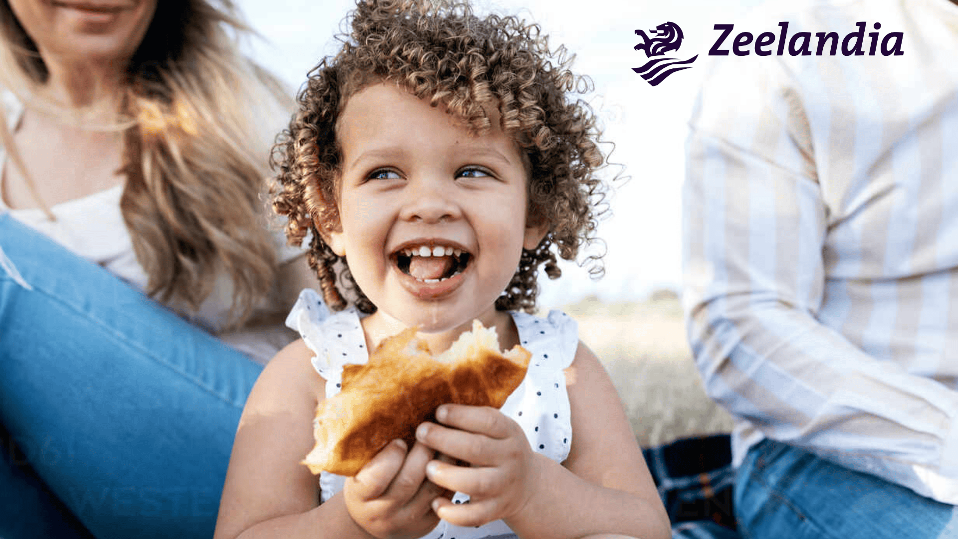 Ein lächelndes Kind mit lockigem Haar hält ein Gebäck und sitzt im Freien zwischen zwei Erwachsenen. Das Zeelandia-Logo ist in der oberen rechten Ecke sichtbar.
