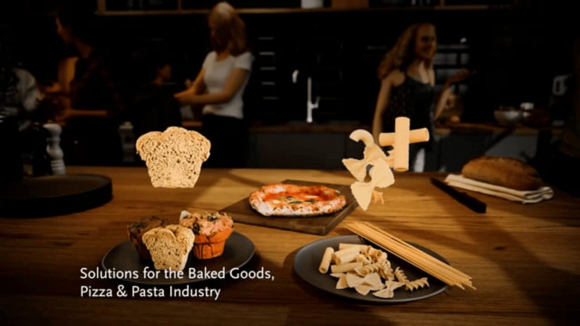 A wooden table with bread, muffins, pizza, and various pasta shapes. Three labeled bread, pizza, and pasta icons float above the food. People stand and talk in the blurred background. Text reads: Solutions for the Baked Goods, Pizza & Pasta Industry.