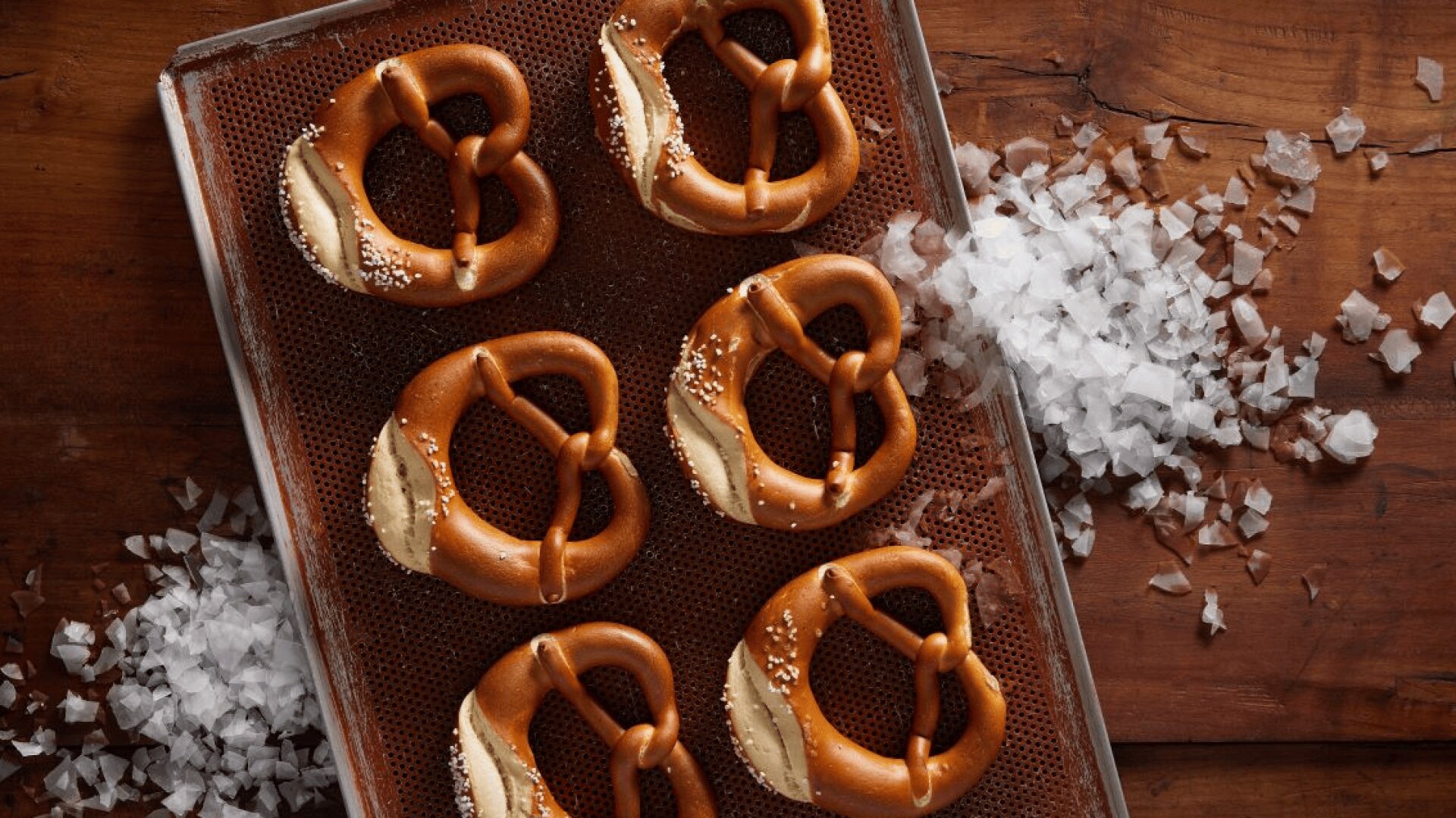 Auf einem Backblech liegen sechs goldbraune, weiche, mit grobem Salz bestreute Brezeln, die auf einer mit Meersalz bestreuten Holzunterlage ruhen.