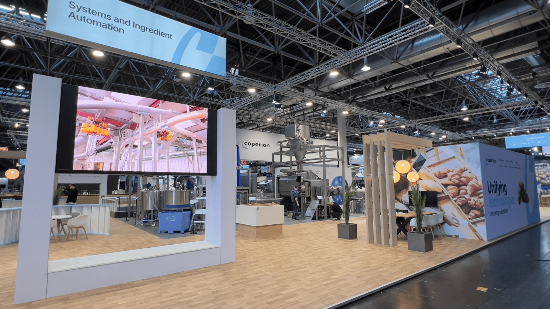 Messestand für System- und Zutatenautomatisierung mit Maschinen, einem großen Bildschirm mit Industrieausrüstung, Sitzbereichen und einer Ausstellung mit Bildern von Backwaren unter heller Deckenbeleuchtung.