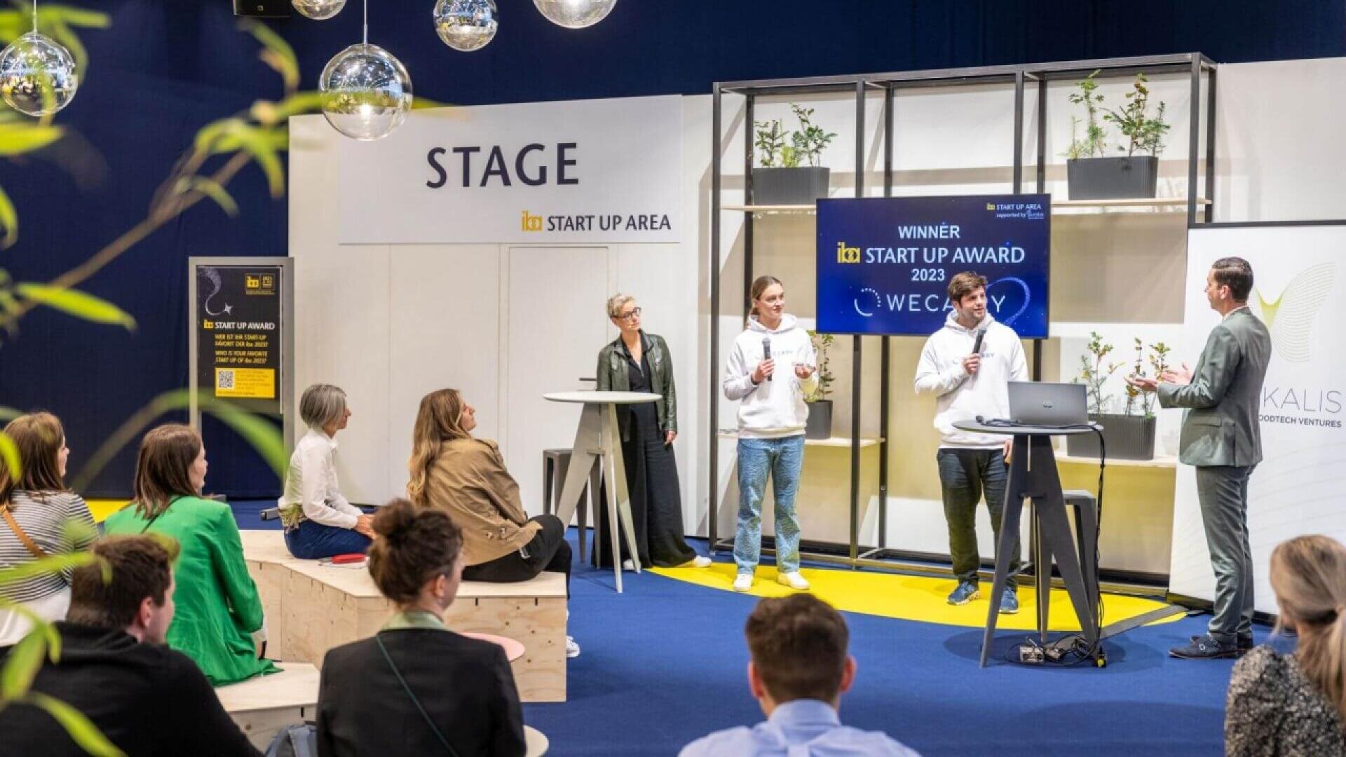 A group of people sit and watch as two presenters stand on a stage with microphones. A large sign reads “Winner Start Up Award 2023.” Plants and decorative lights are visible. An audience faces the stage.