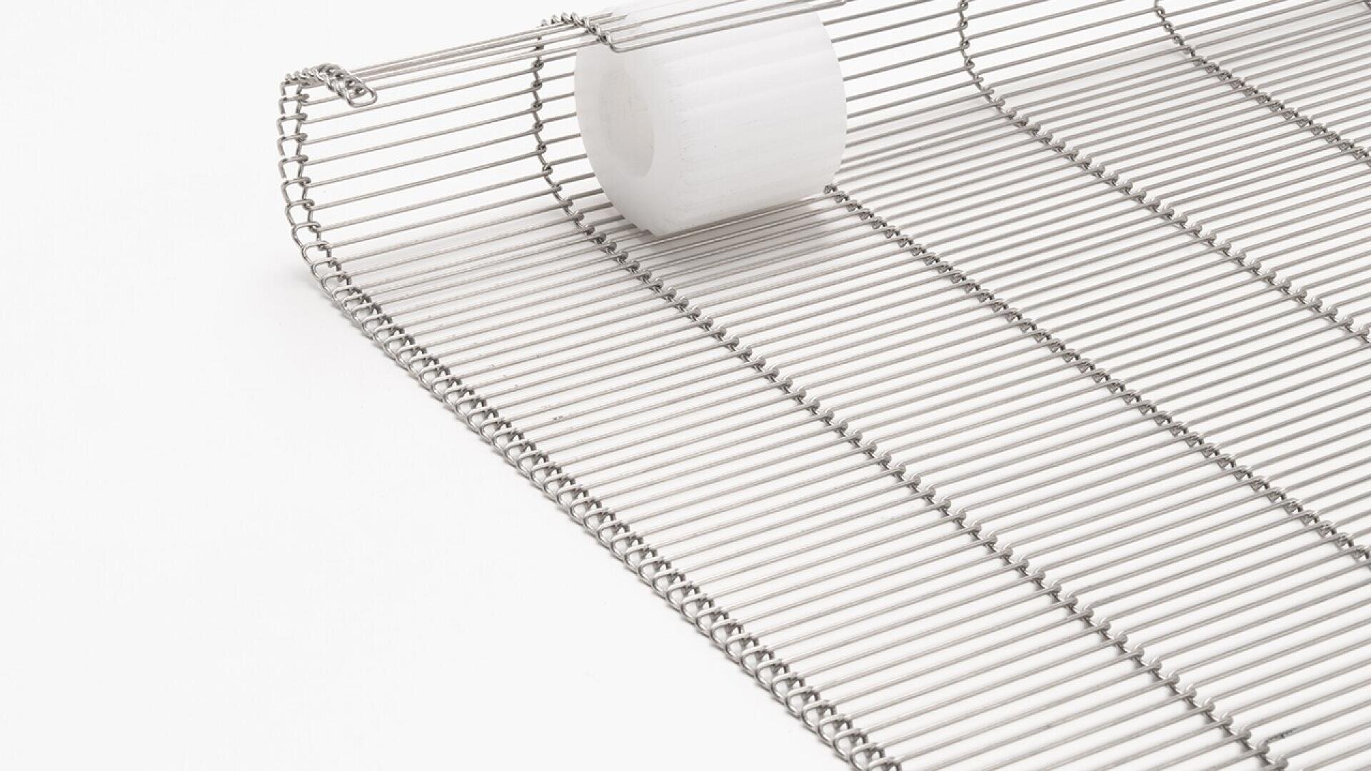 Nahaufnahme eines Förderbands aus Metalldraht mit gleichmäßig verteilten Stäben und einer weißen Kunststoffrolle oben drauf, alles vor einem schlichten weißen Hintergrund.