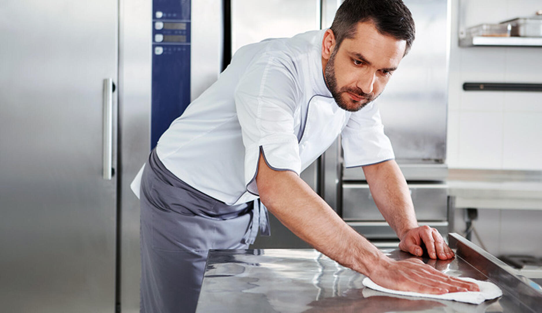 Ein Koch in weißer Uniform und grauer Schürze wischt eine Edelstahltheke in einer professionellen Küche ab und konzentriert sich auf die Reinigung der Oberfläche.