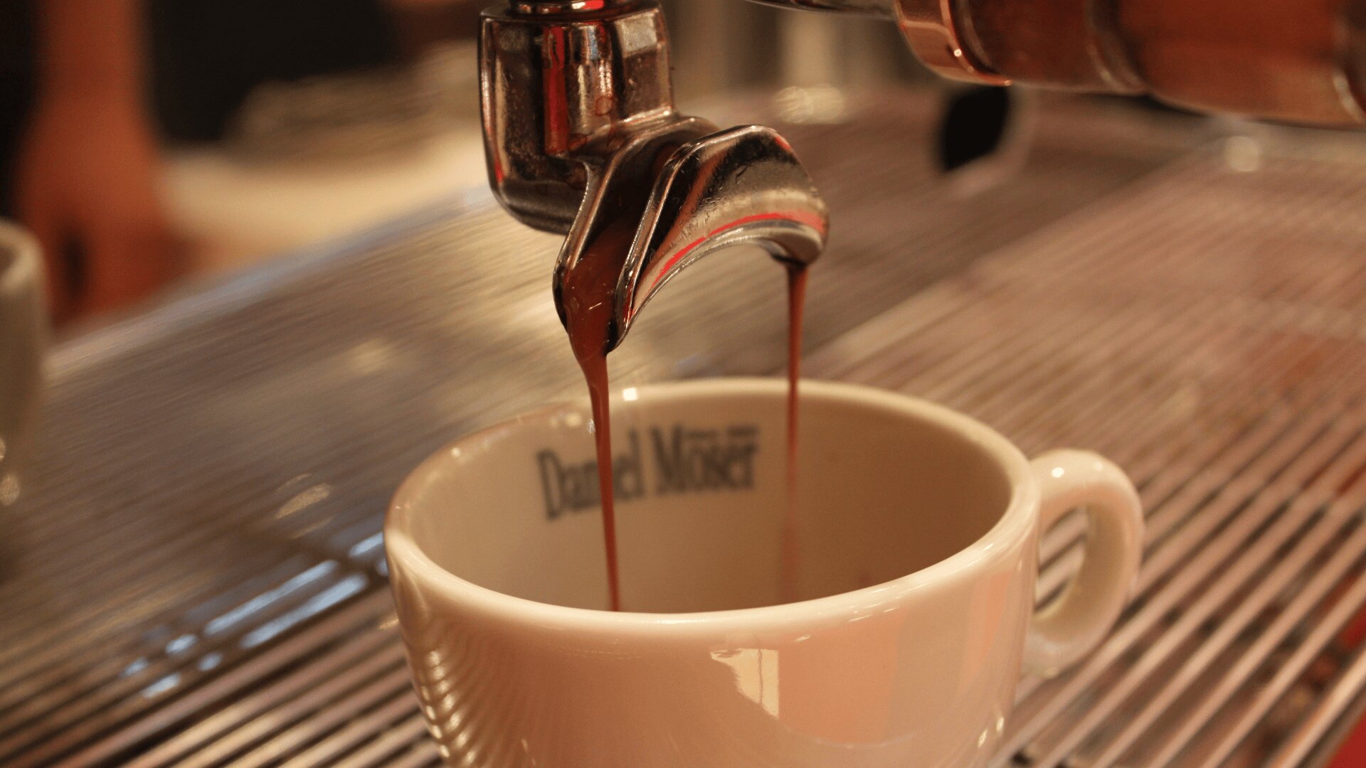 Espresso fließt aus dem Siebträger einer Maschine in eine weiße Keramiktasse, die auf einer Metallabtropfschale steht. Am Rand der Tasse befindet sich eine verschwommene Beschriftung.