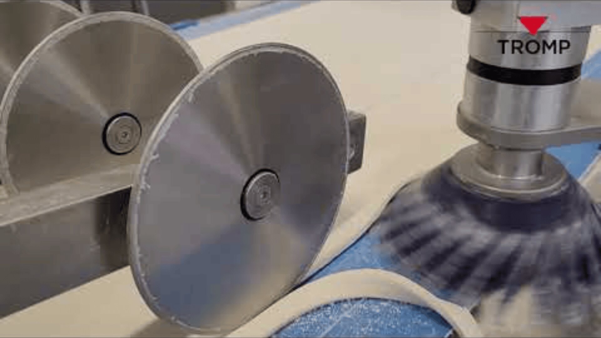 Nahaufnahme einer Lebensmittelverarbeitungsmaschine, die Teig mit großen Metallkreisklingen und einer rotierenden Bürste auf einem blauen Förderband in Streifen schneidet. Das TROMP-Logo ist in der oberen rechten Ecke sichtbar.