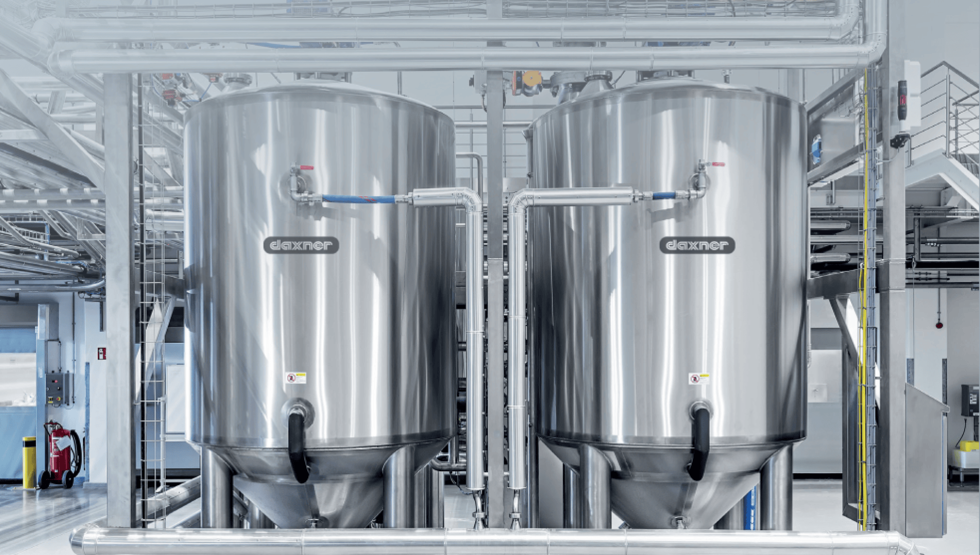 Zwei große Edelstahltanks mit Dexner-Etiketten stehen nebeneinander in einer Industrieanlage. Im Hintergrund sind Metallrohre, Plattformen und Sicherheitsgeländer zu sehen.