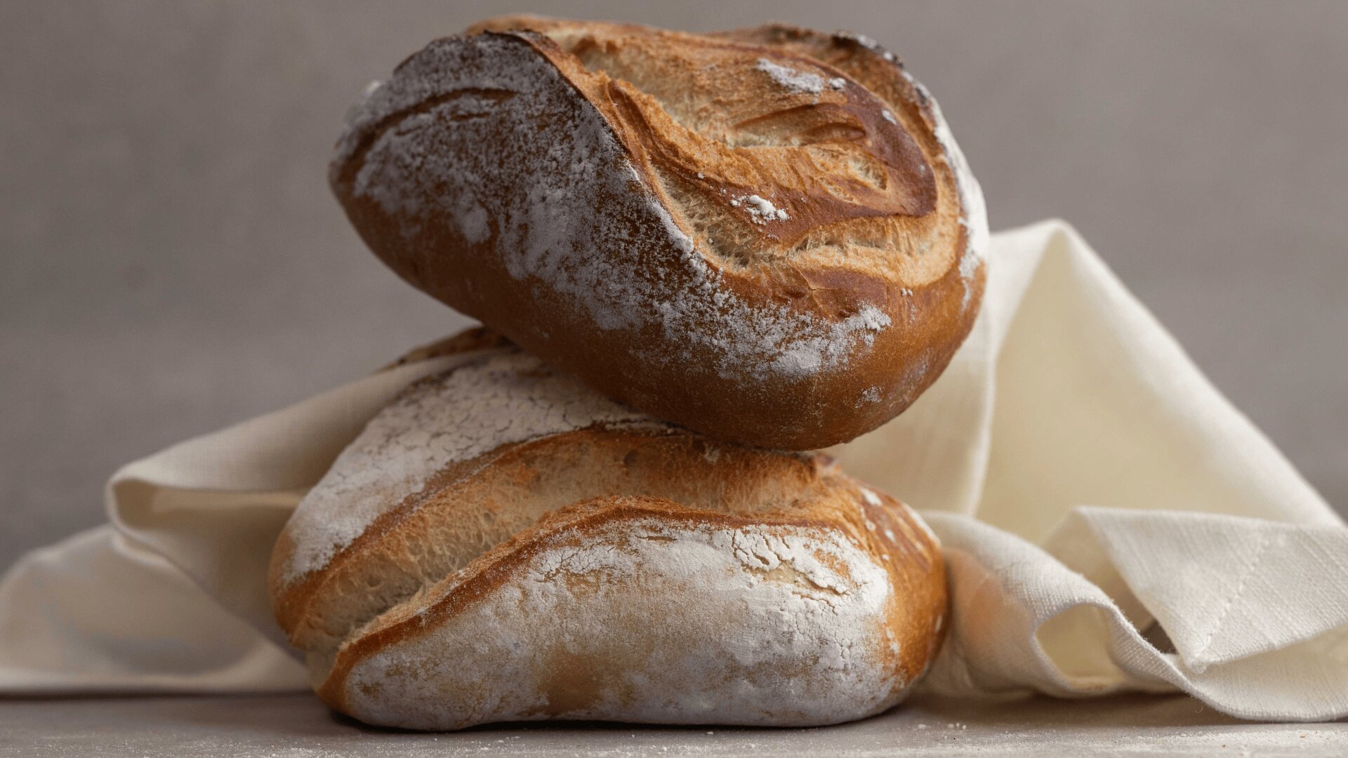 Two rustic loaves of artisan bread rest on a light beige cloth, with a neutral background. The bread has a golden-brown crust and light dusting of flour, showing off its textured surface.