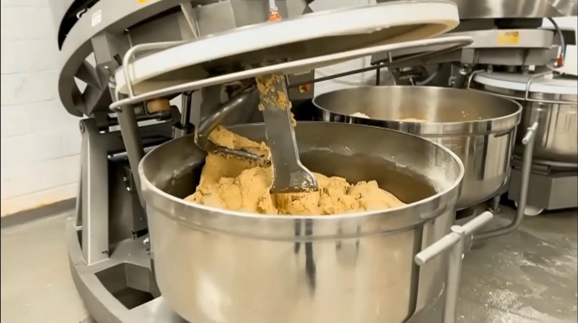 A large industrial mixer with a metal bowl is mixing dough in a commercial kitchen or bakery setting. The mixing paddle is coated in thick, light brown dough.