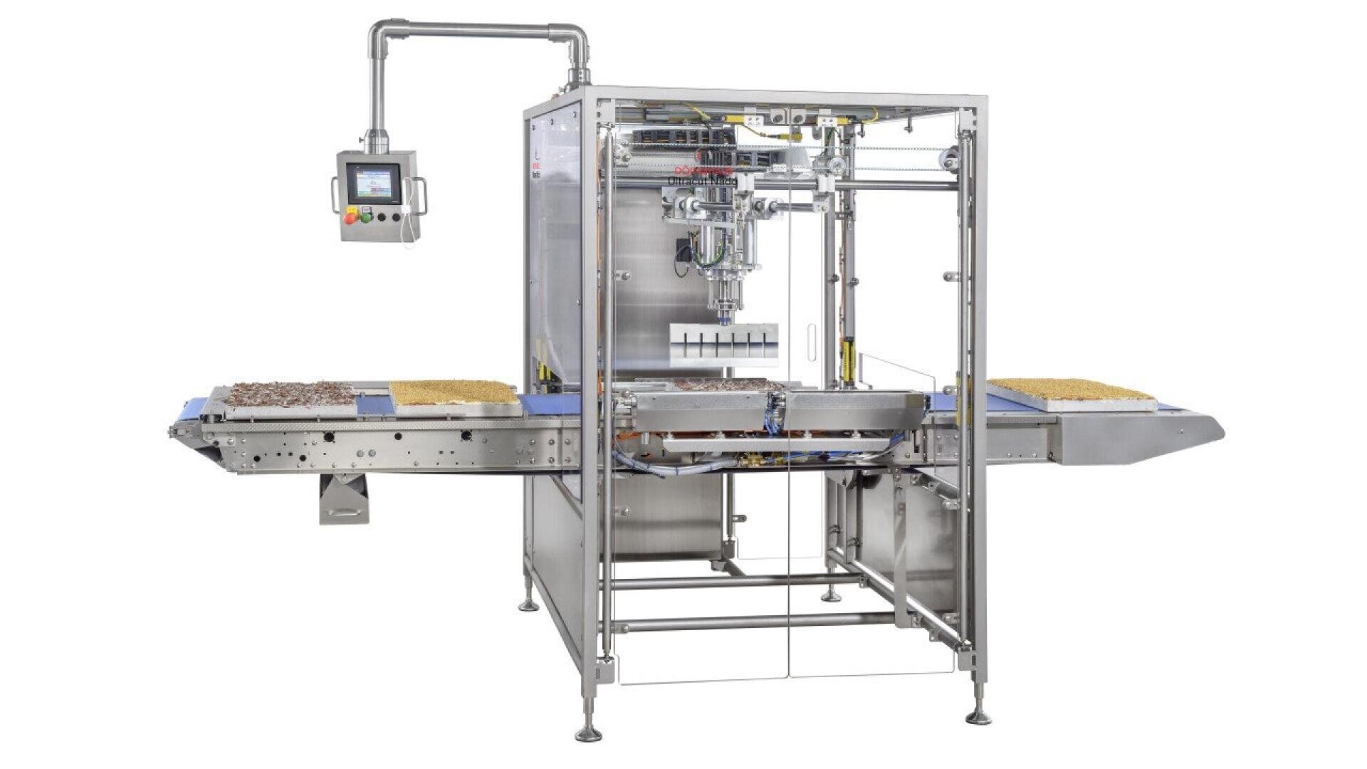 A stainless steel industrial food processing machine with conveyor belts carrying food items on both sides; control panel attached to the left side.