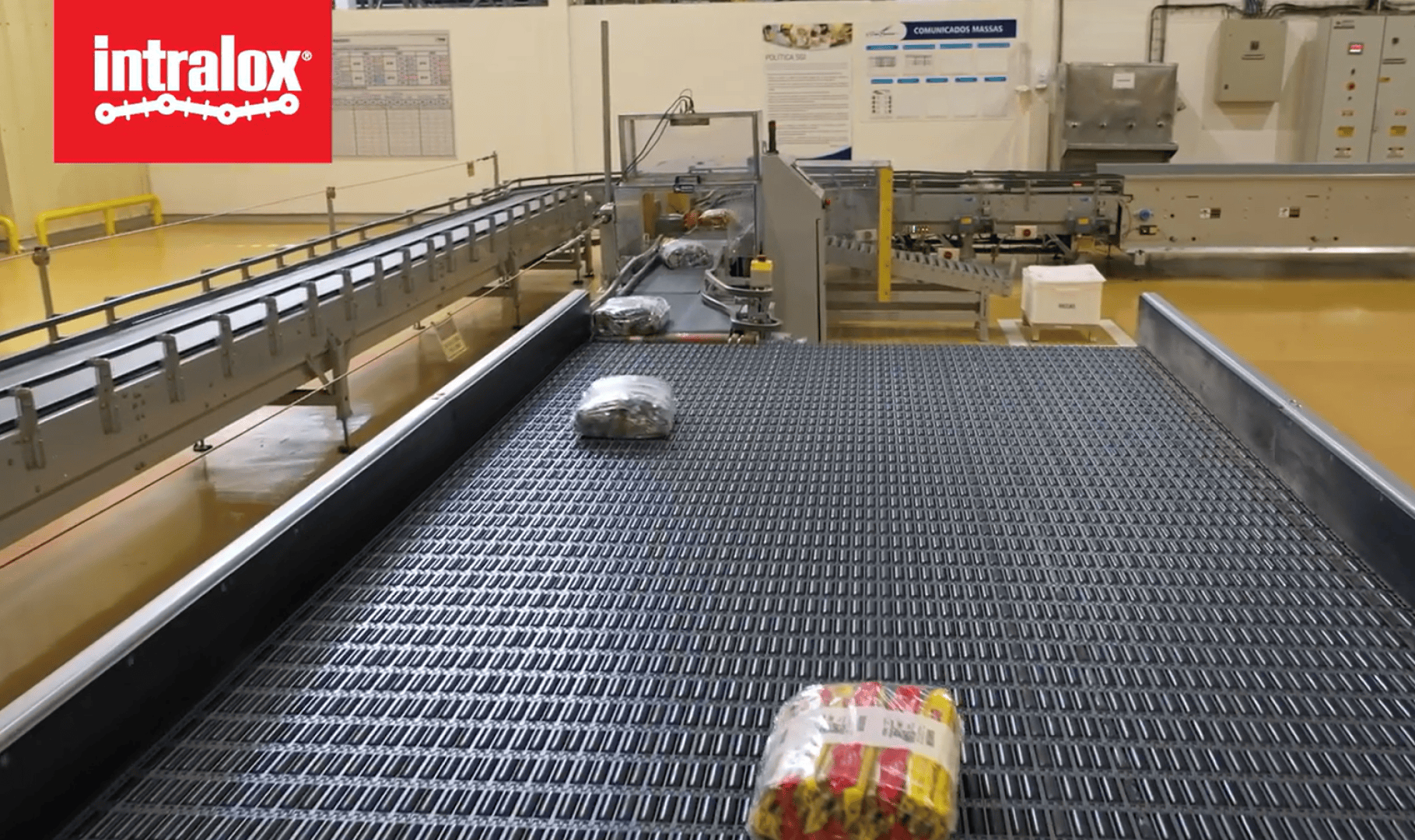 A conveyor system in a factory moves packaged products along black interlocking belts. The Intralox logo appears in the top left corner. The background shows machinery, safety rails, and instruction posters on the wall.