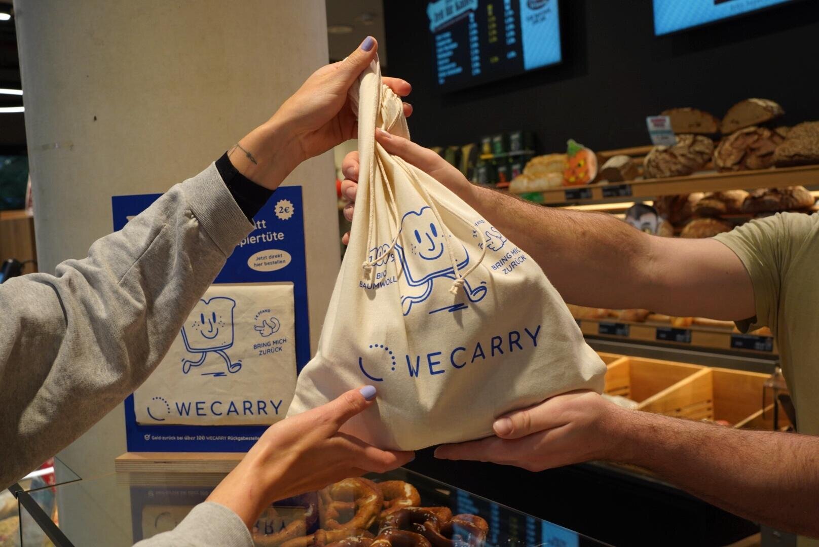 A person hands a cloth bag with the WeCarry logo and a cartoon slice of bread to another person in a bakery, with pretzels and bread displayed in the background.