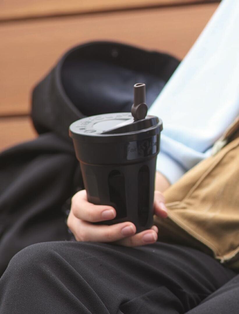 Eine Person hält einen schwarzen Mehrwegbecher mit Deckel und Strohhalm und sitzt im Freien auf einer Bank. Der Becher wird in einer Hand gehalten, und ein Teil der Freizeitkleidung ist sichtbar.