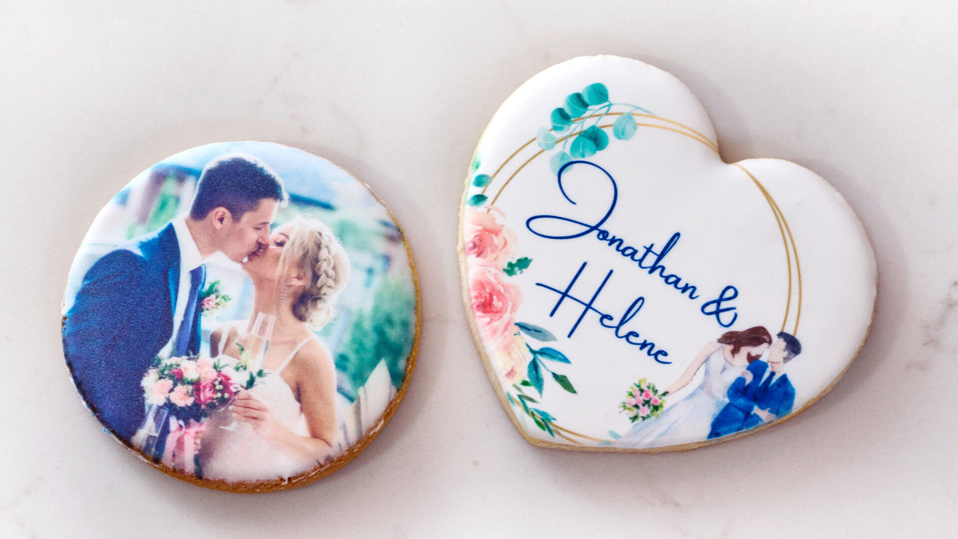 Zwei verzierte Kekse: ein runder mit einem Foto eines sich küssenden Brautpaares und ein herzförmiger mit Blumenmuster und den Namen Jonathan & Helene sowie einem kleinen Bild des Paares.