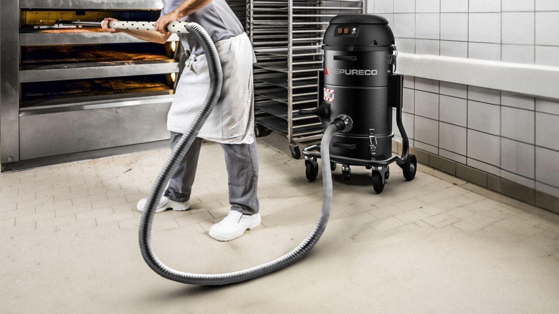 Eine Person in weißen Hosen, Schuhen und einer Schürze verwendet einen großen Industriestaubsauger, um den Fliesenboden in einer Großküche oder Bäckerei zu reinigen. Im Hintergrund sind ein Ofen und Metallgestelle zu sehen.