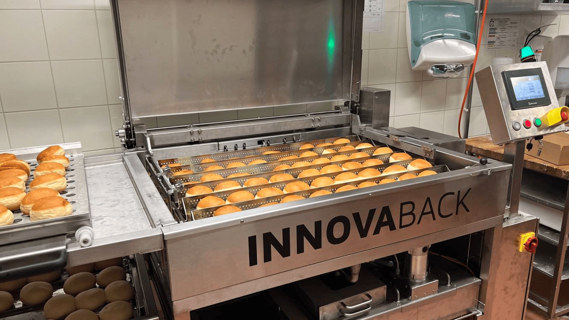 Eine Bäckereimaschine mit der Aufschrift INNOVABACK frittiert Reihen von runden Brötchen. Einige fertige Brötchen liegen auf einem Tablett neben der Maschine. Im Hintergrund sind ein Bedienfeld und ein an der Wand montierter Handschuhspender zu sehen.