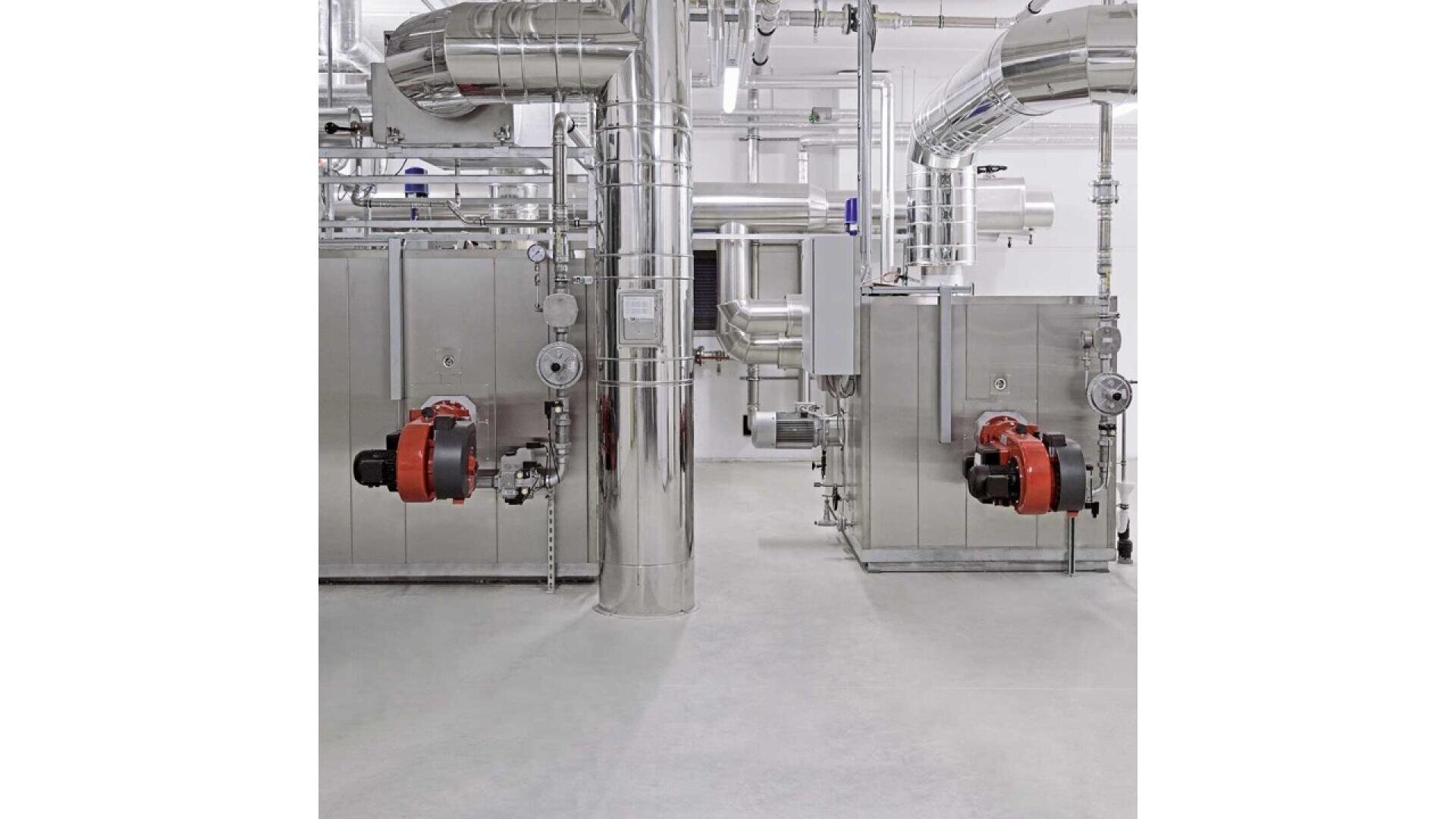Two large industrial boilers with red burner units are installed in a clean, bright mechanical room. Shiny metal pipes and ducts run overhead against white walls and a smooth gray floor.