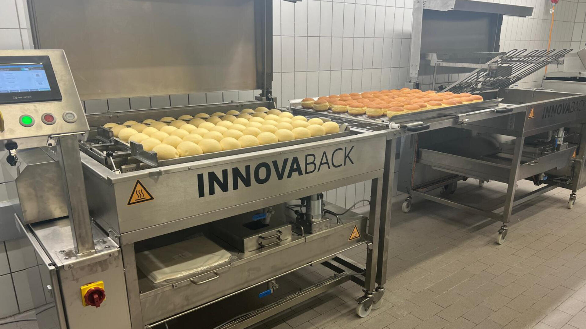 Teigbällchen und goldbraune Brötchen sind auf einer industriellen Bäckereimaschine von INNOVABACK in einer gefliesten Bäckereiumgebung aufgereiht, links ist ein Bedienfeld sichtbar.