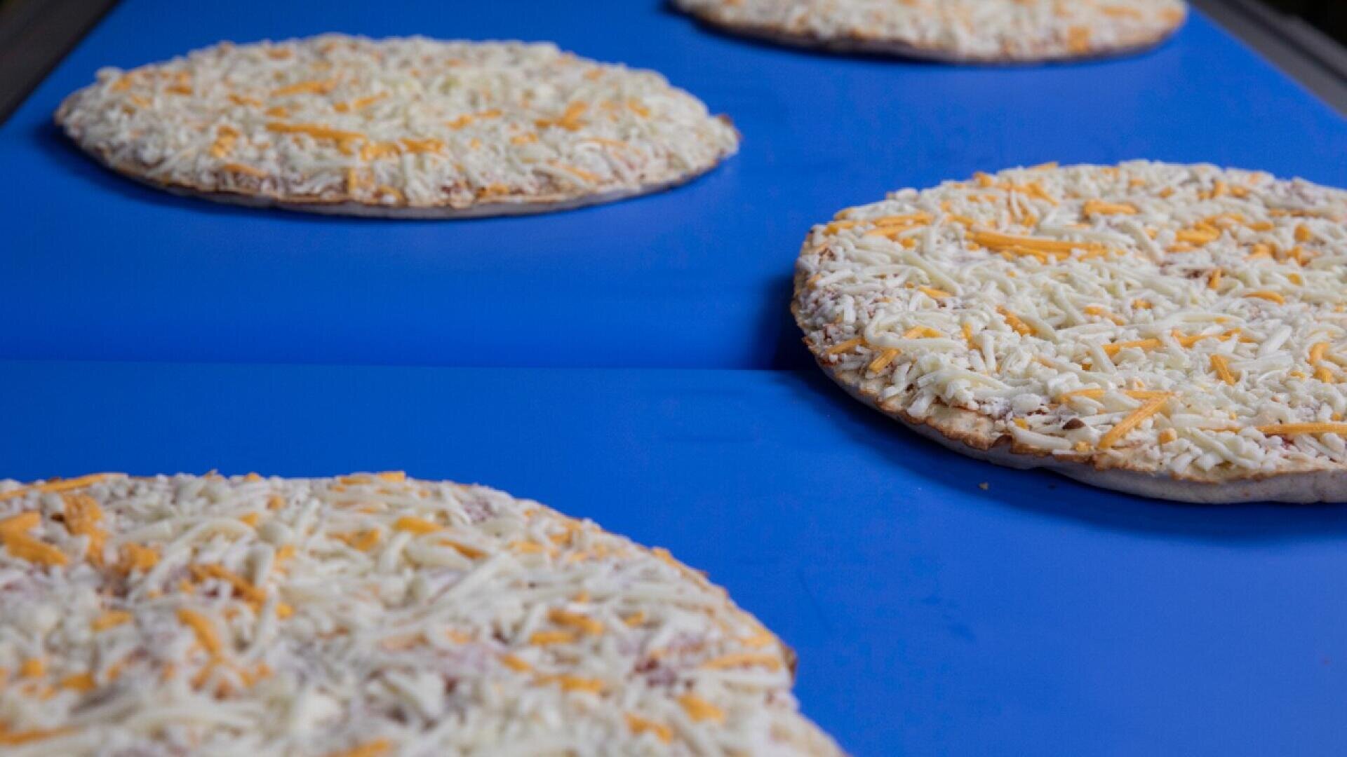 Vier ungebackene Tiefkühlpizzen mit geriebenem Käse liegen in einer Reihe auf einem blauen Förderband, wahrscheinlich in einer Lebensmittelproduktions- oder -verpackungsanlage.